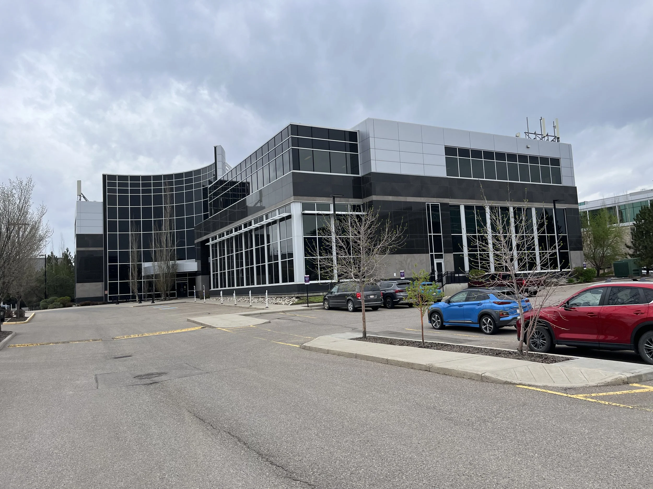 A modern building with black and white glass windows, a parking lot with several cars, leafless trees, and a cloudy sky.