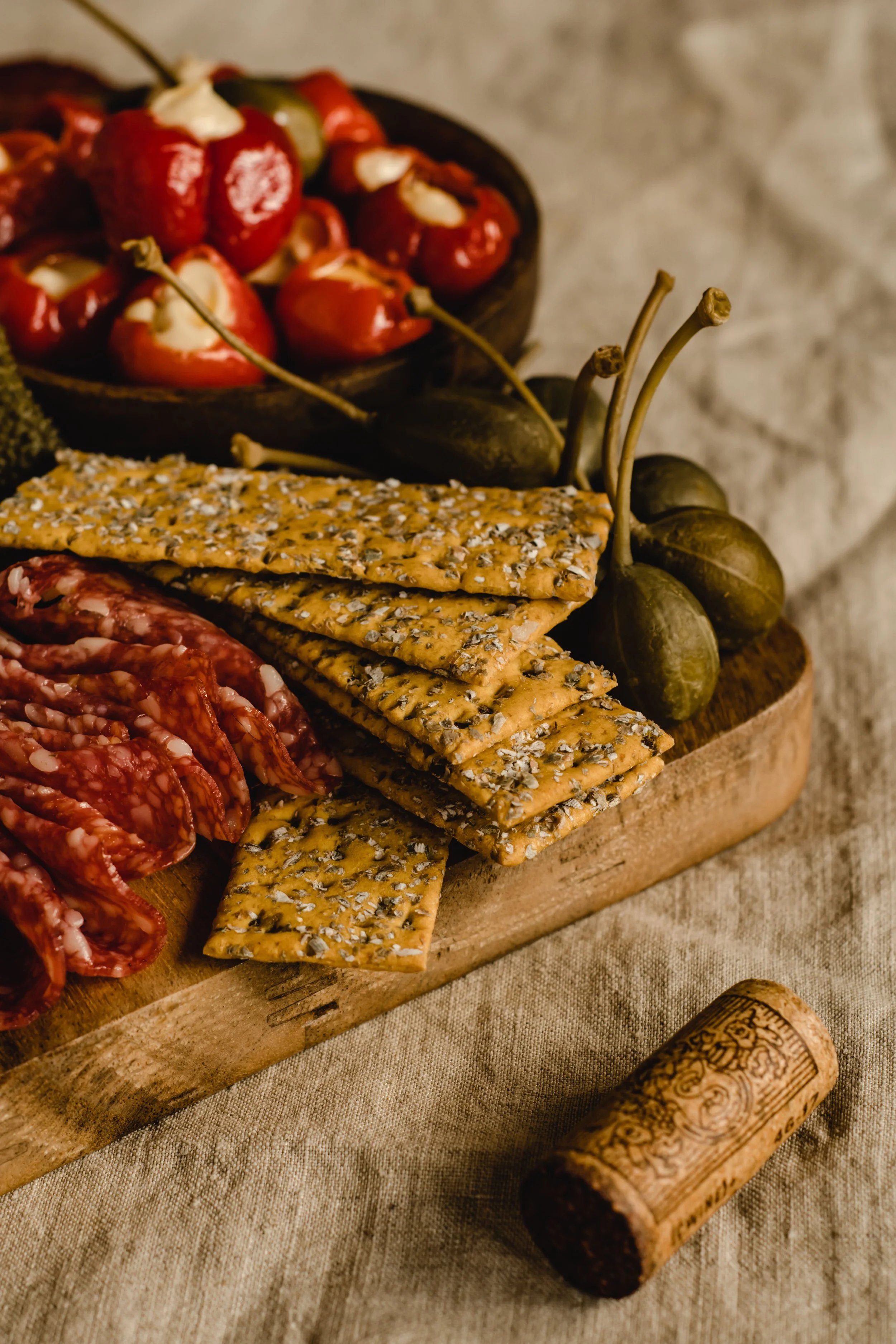 A wooden serving board with dried salami, sesame seed crackers, green and red peppers, and grapes, set on beige fabric with wine cork decor.