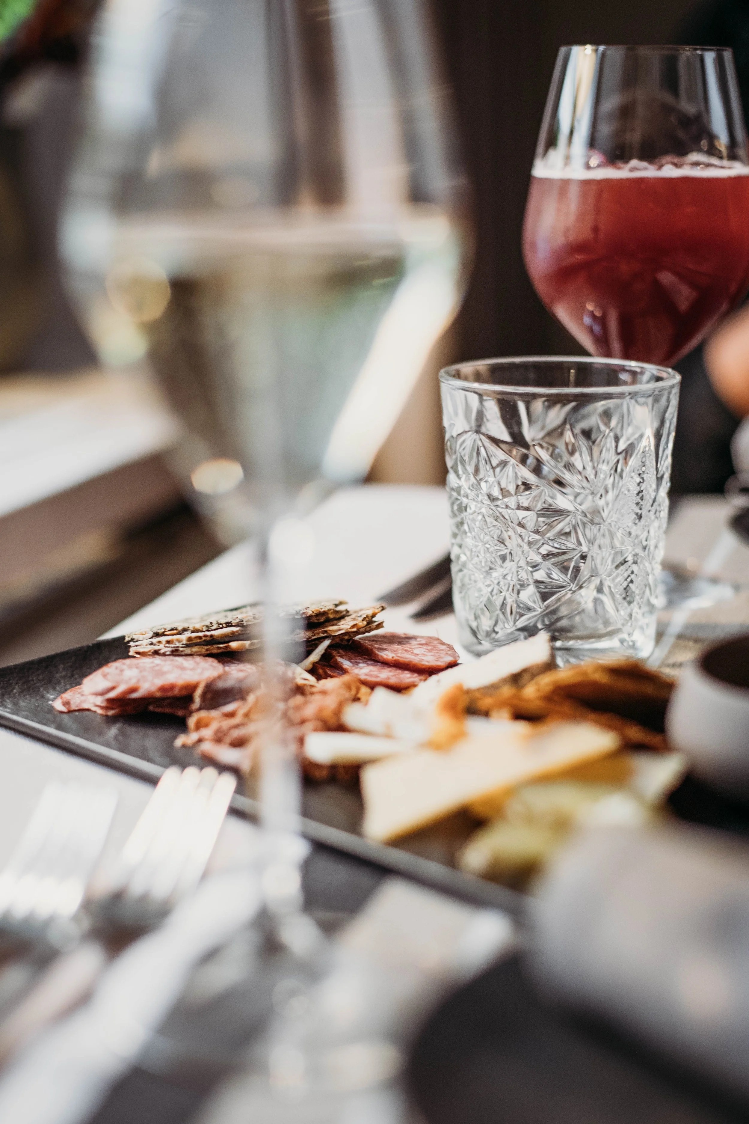 A close-up of a charcuterie board with sliced meats and cheeses, with glassware including a wine glass, a water glass, and a crystal glass, set on a table at a social gathering or restaurant.