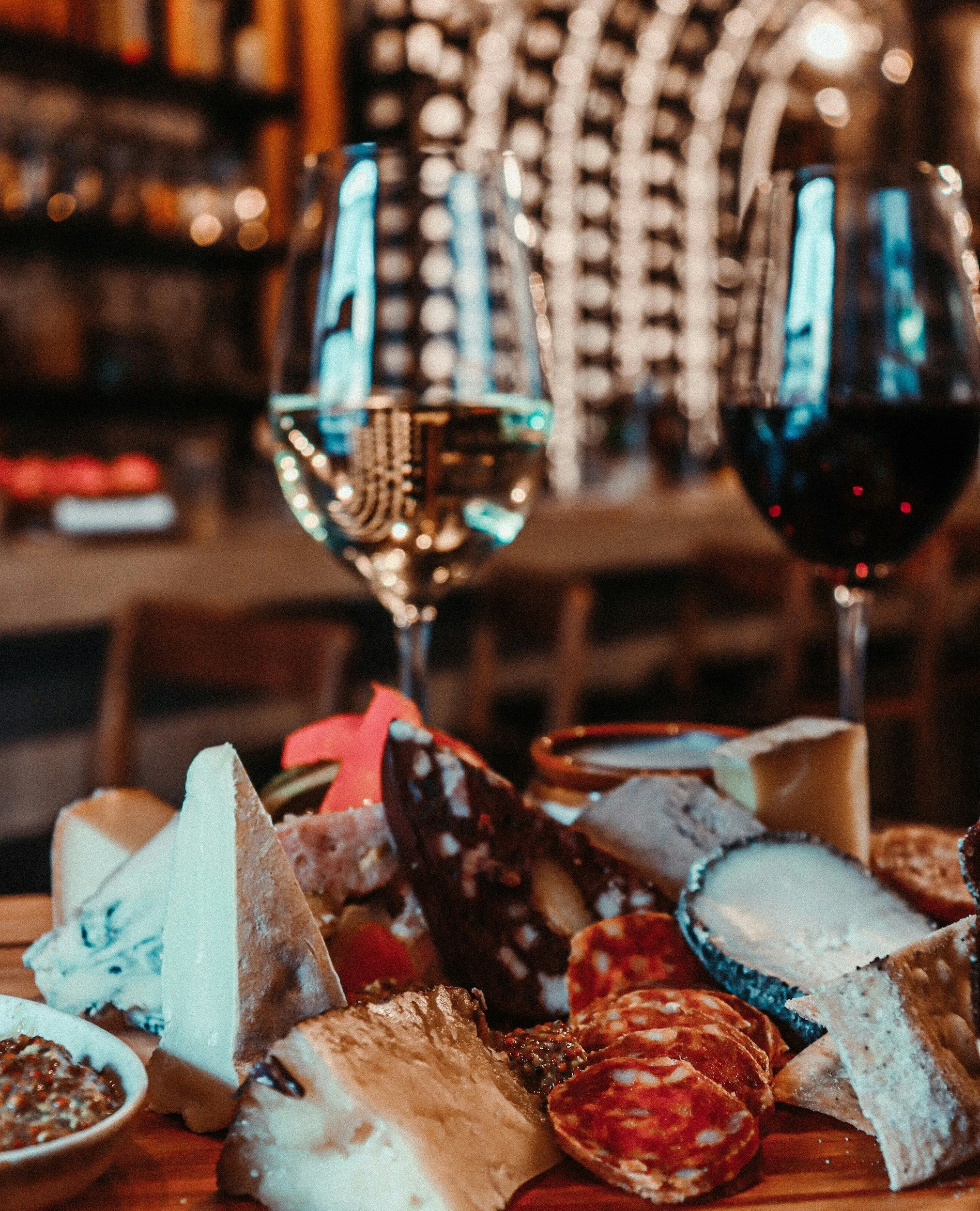 Close-up of assorted cured meats, cheeses, and spreads on a wooden platter with a blurred background of shelves and bokeh lights.