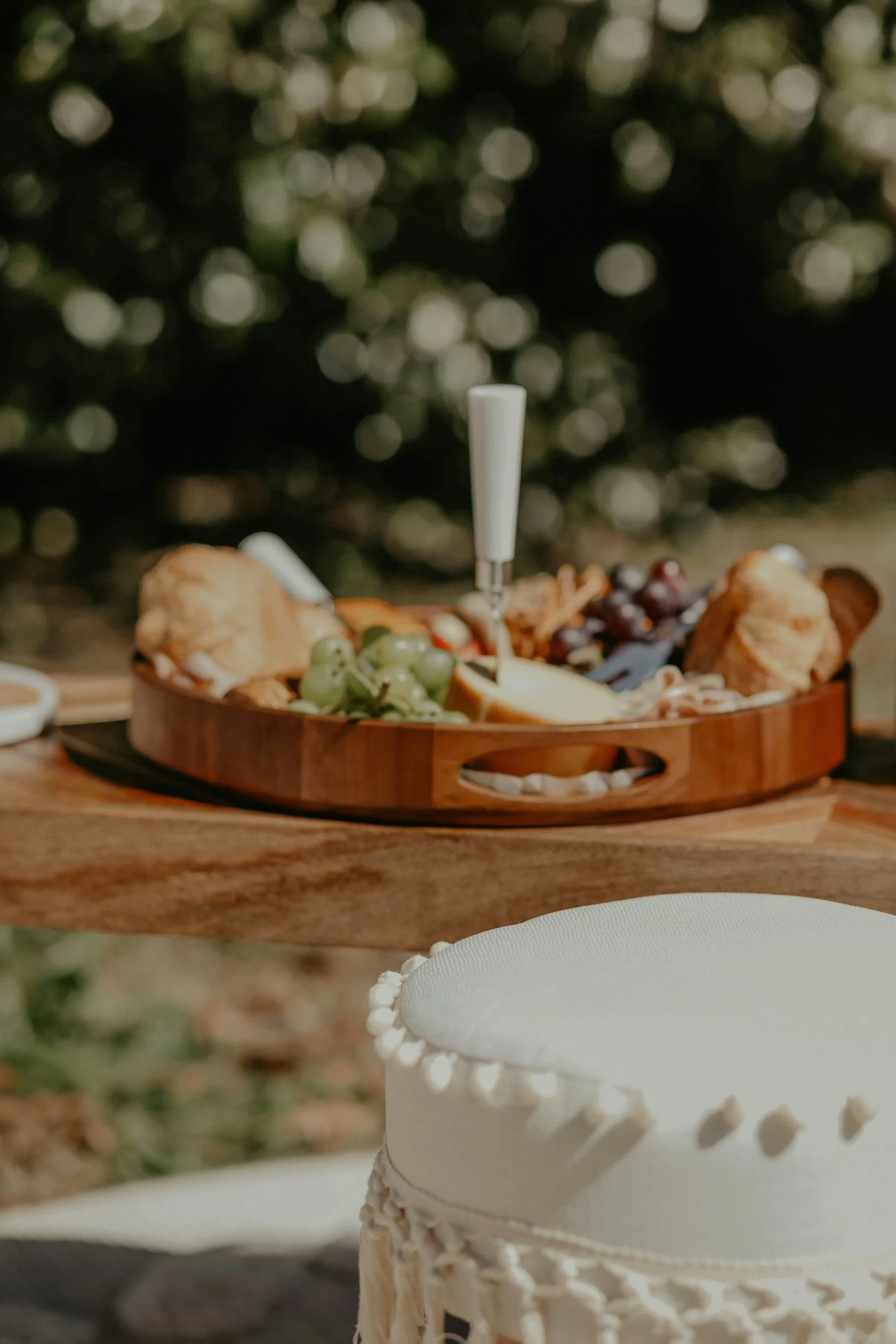 Close-up of a white wedding cake with decorative piping and a white hat-shaped figure on top, set on a table outdoors with a blurred background of greenery and bokeh lights.