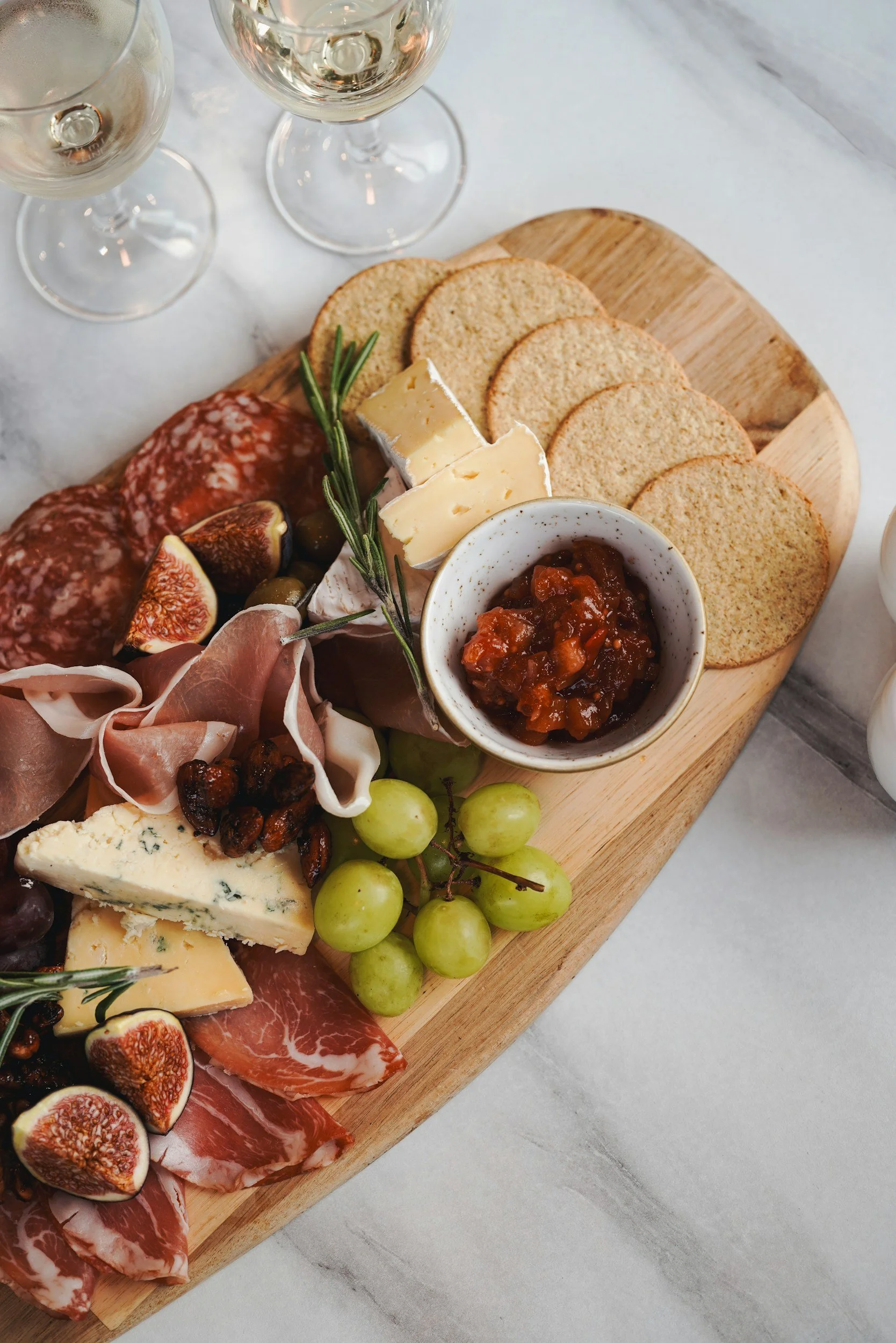 Charcuterie board with cheese, crackers, grapes, figs, cured meats, and a small bowl of fruit chutney, set on a light marble surface.