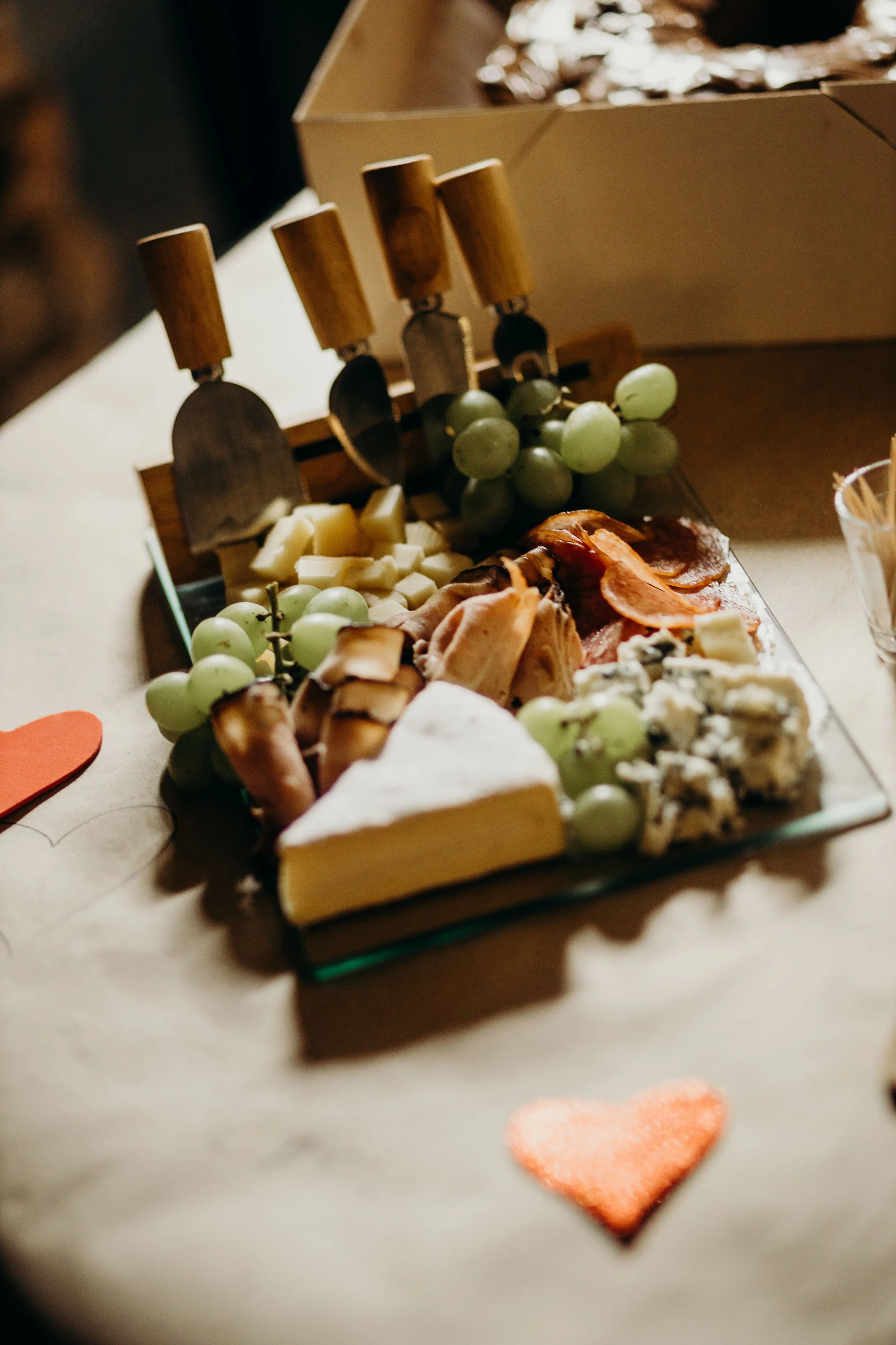 A cheese and charcuterie board with grapes, different cheeses, sliced cured meats, and a heart-shaped strawberry, with a cardboard box in the background.