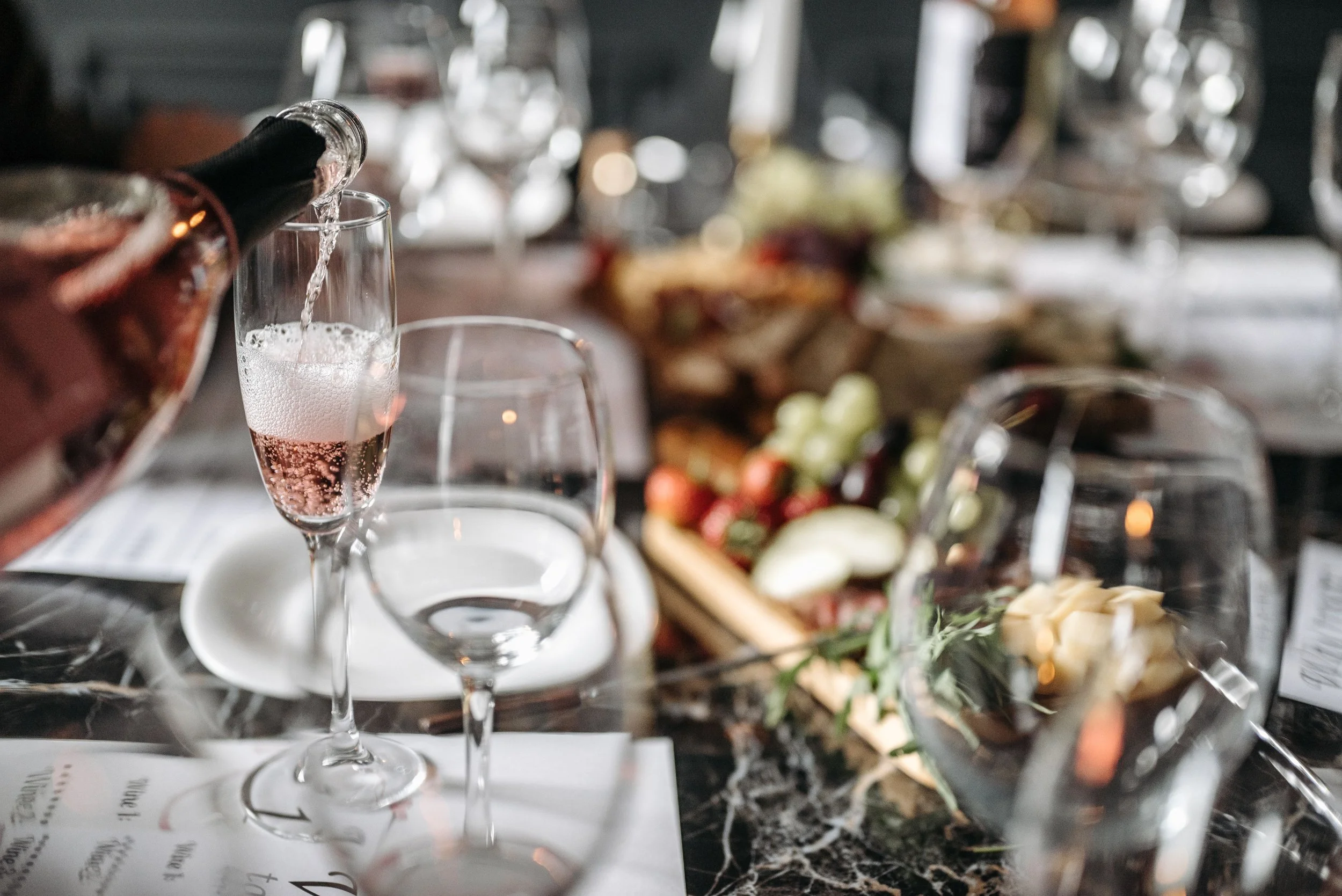 A glass of sparkling rose wine being poured at a social gathering with a table of food and drinks in the background