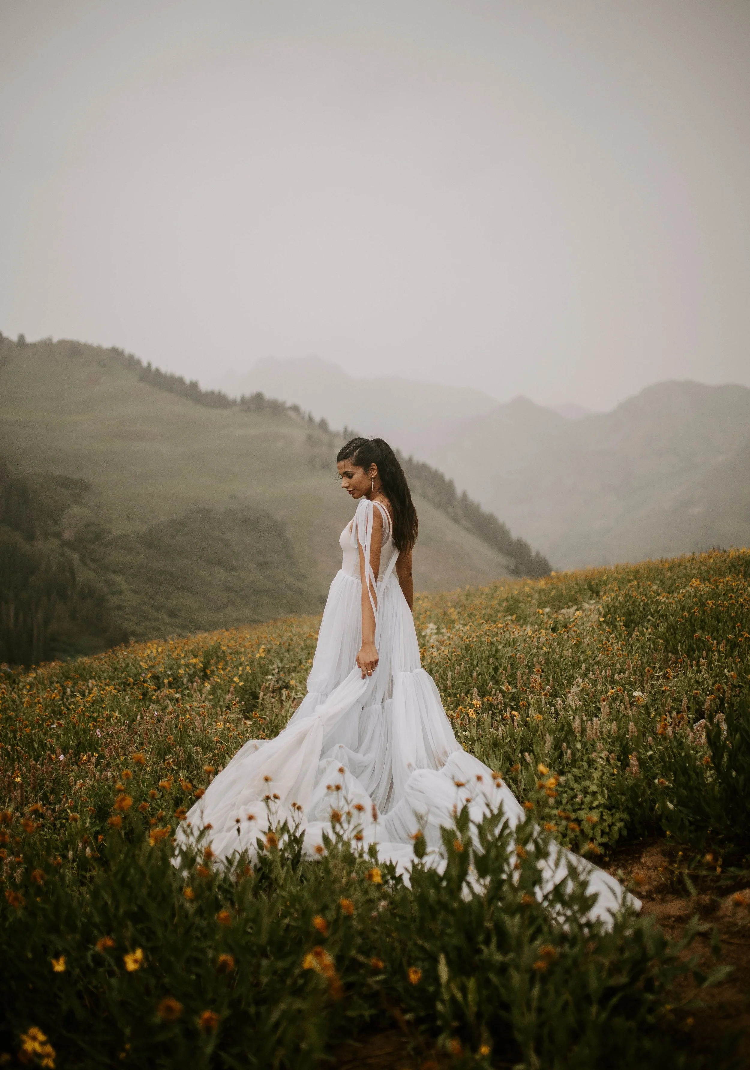 A woman in a white dress with a train standing in a field of orange flowers with green hills and mountains in the background, under a gray sky.