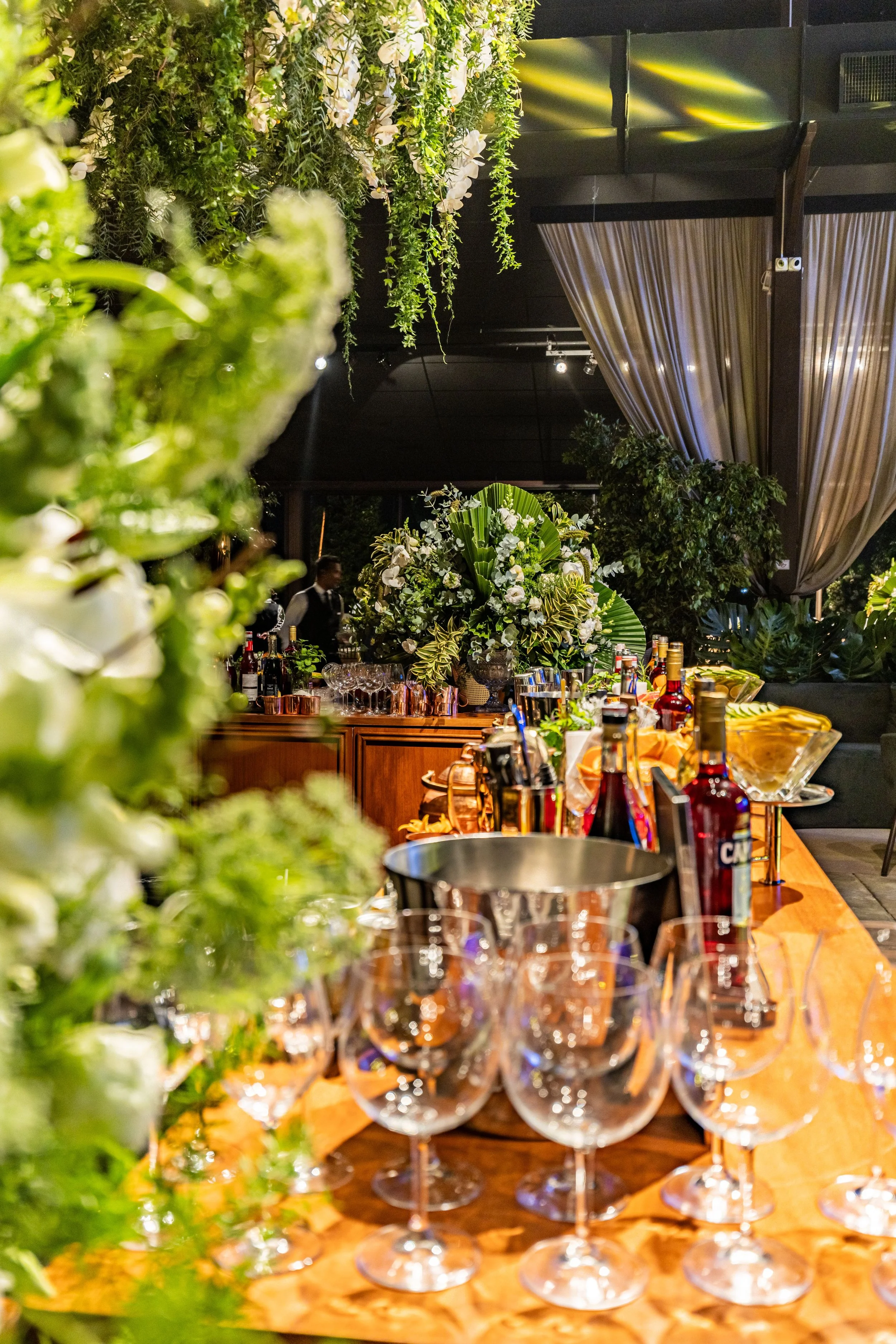 A bar or event space decorated with lush green foliage, white flowers, and a large floral arrangement on a wooden table. The table is set with empty wine glasses, bottles of alcohol, and barware. Curtains hang in the background, and a waiter can be seen in the distance.