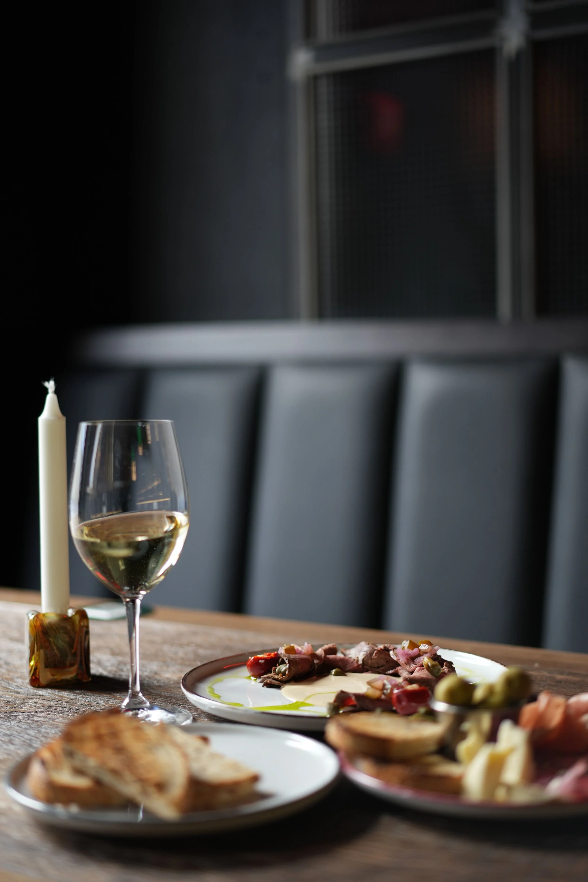 A dining table set with a glass of white wine, a plate of meat with vegetables, bread, and a lit white candle, with a dark background featuring chairs and metal shelves.