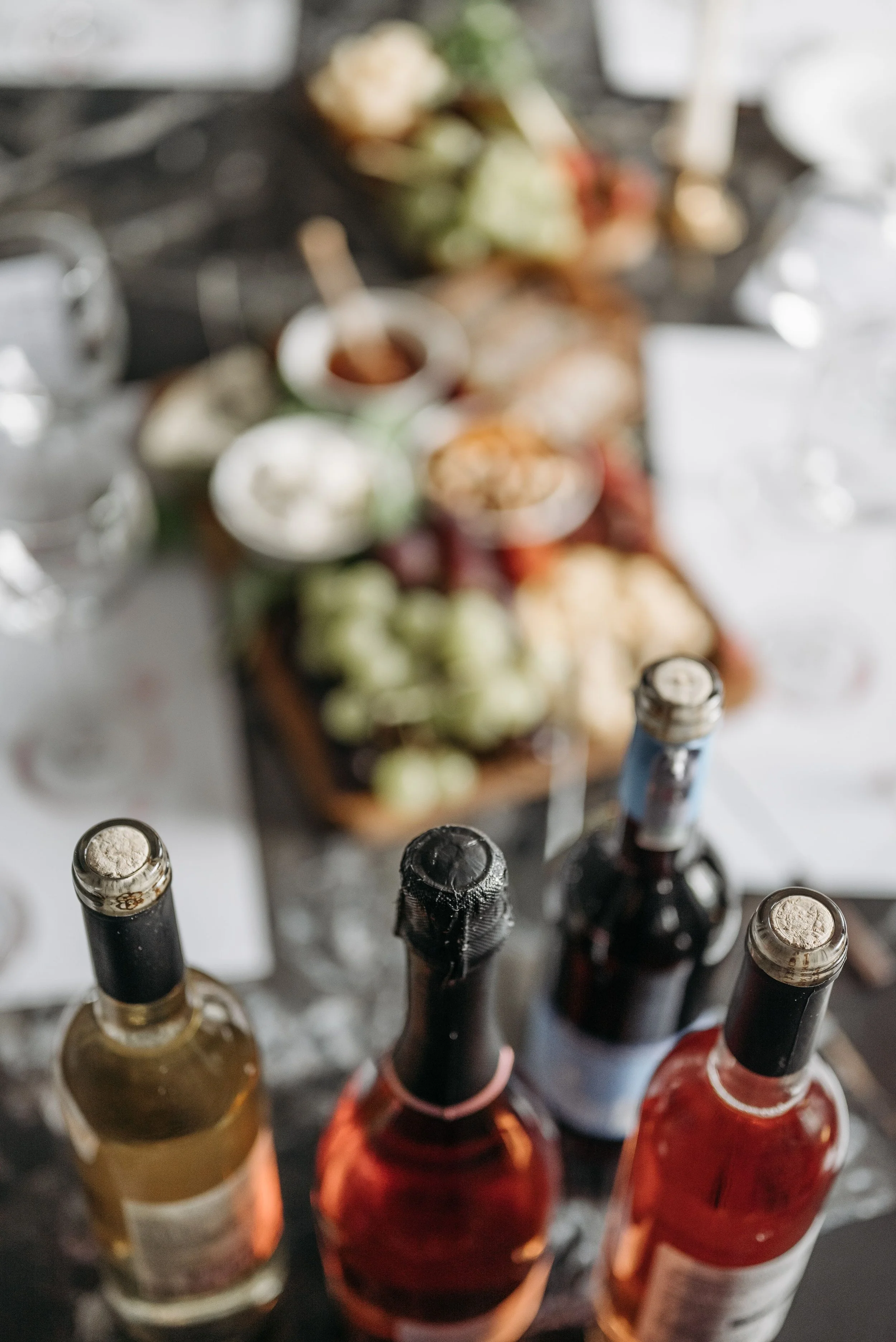A variety of alcohol bottles with cork or cap tops seen from a top-down perspective, with a blurred plate of food in the background.