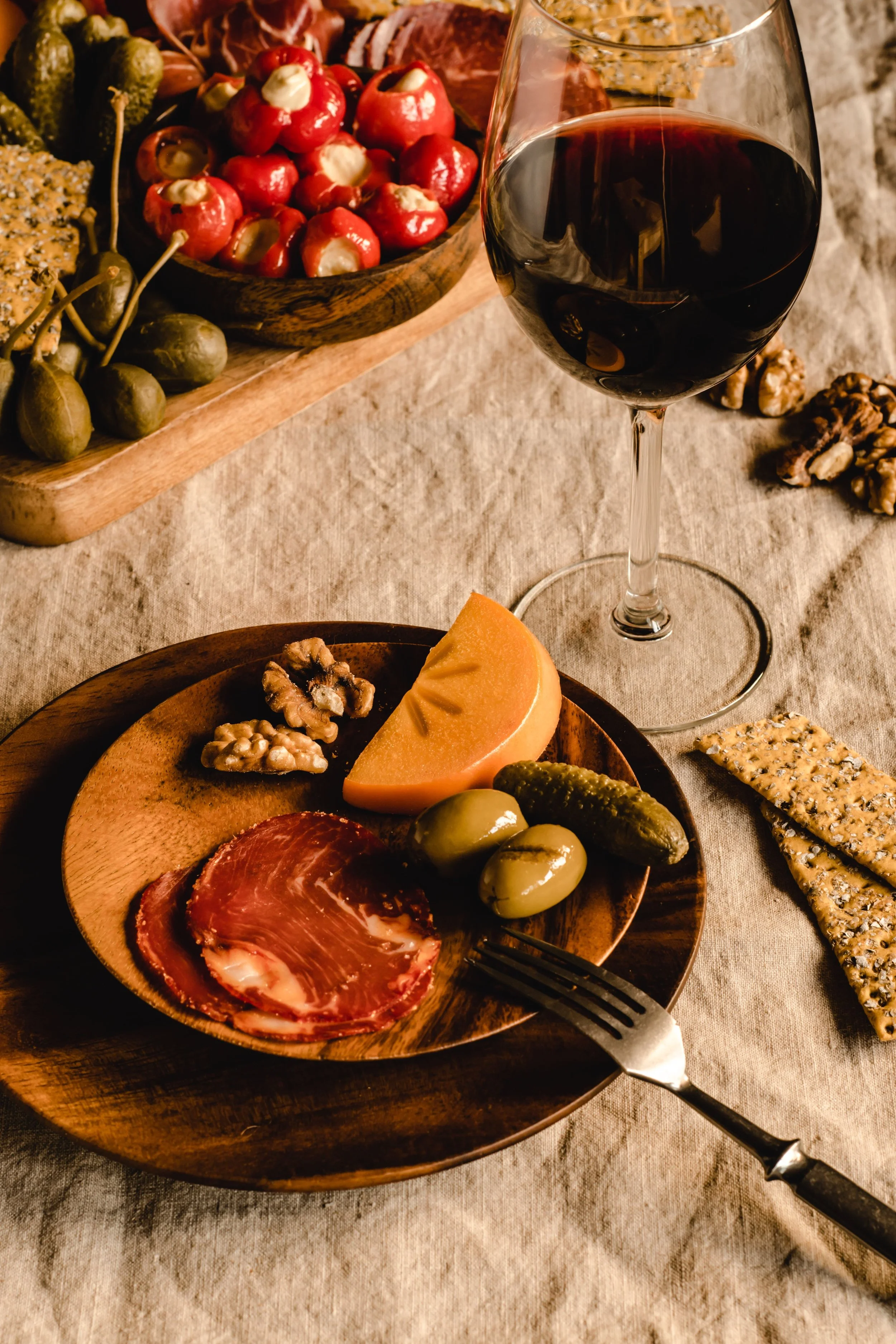 A glass of red wine, a wooden plate with assorted cheeses, nuts, green olives, a slice of cantaloupe, slices of cured meats, and crackers on a table with a textured tablecloth.