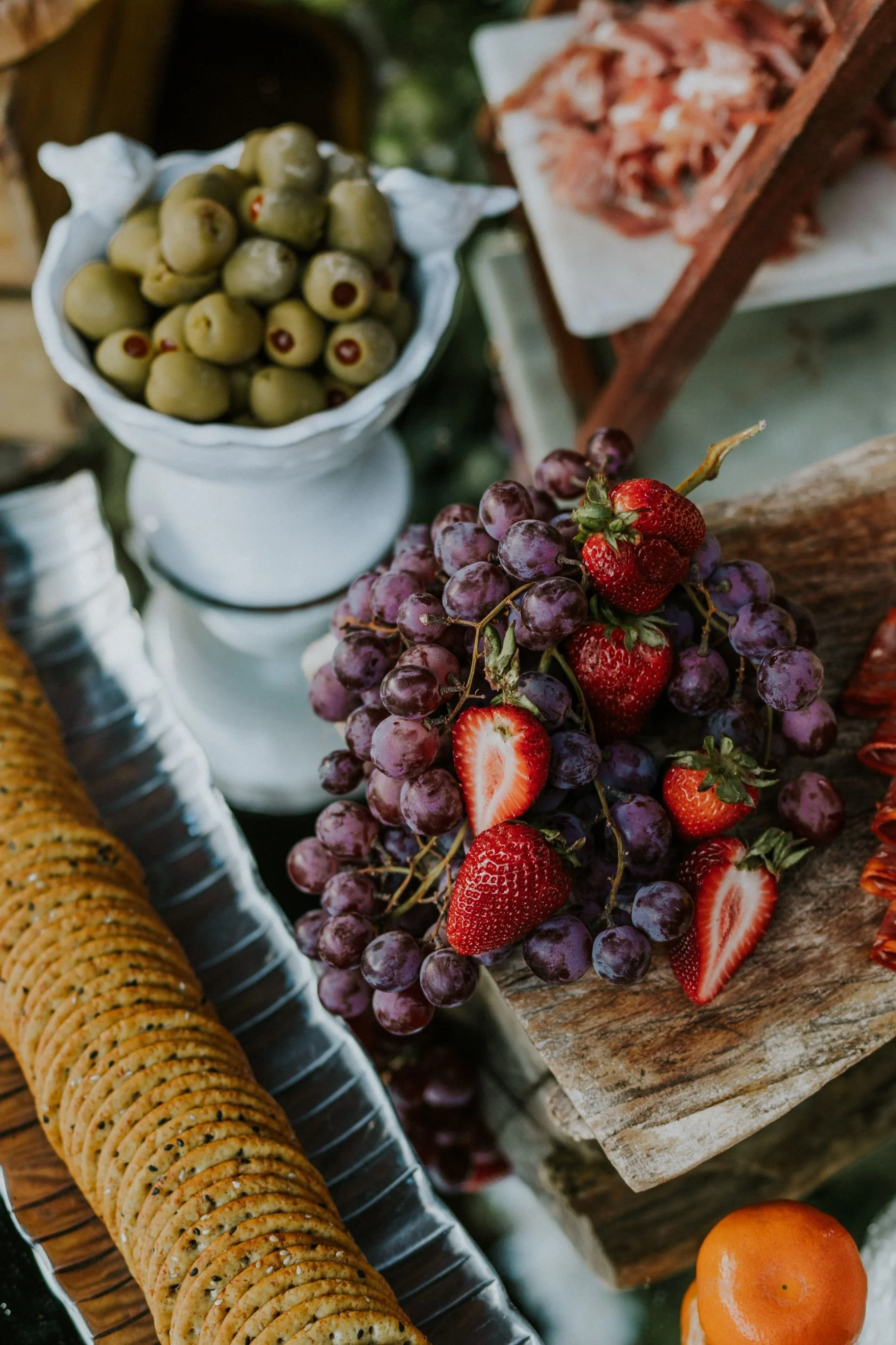 Assorted fresh fruits including green olives in a white bowl, purple grapes with strawberries, and an orange fruit on a rustic wooden surface.