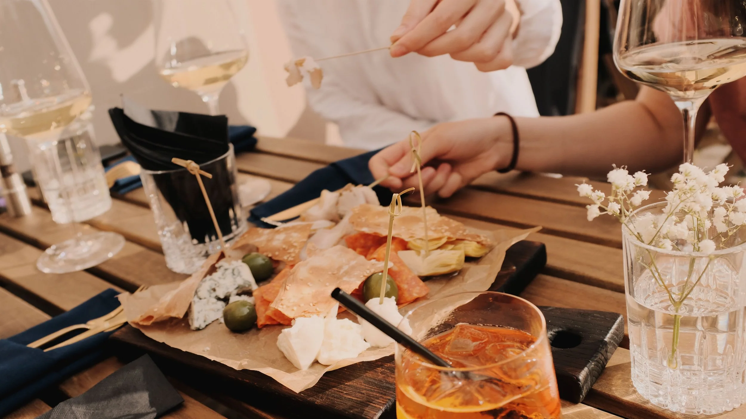 A close-up of a table set for dining, featuring a charcuterie board with various cheeses, cured meats, and green olives. There are three glasses of white wine, a small black bucket with black napkins, and a vase with white flowers. A person is seen reaching for the charcuterie with a toothpick, with a hand visible.