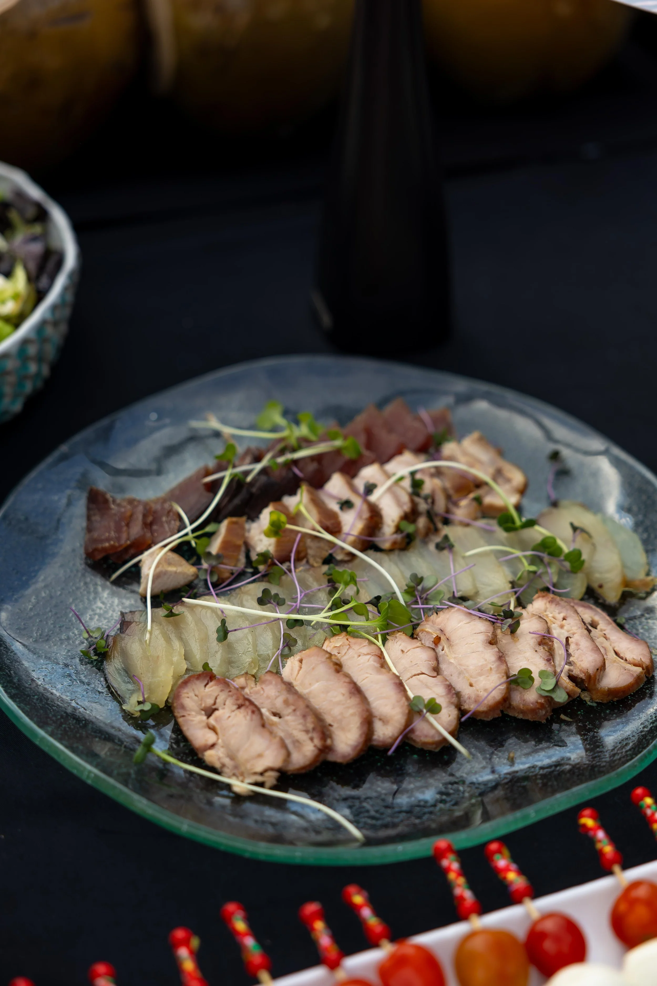Assorted foods including sliced meats, potatoes, and microgreens on a decorative plate, with a dark drink in a black bottle in the background, and red and white candy canes and cherry-topped treats in the foreground.