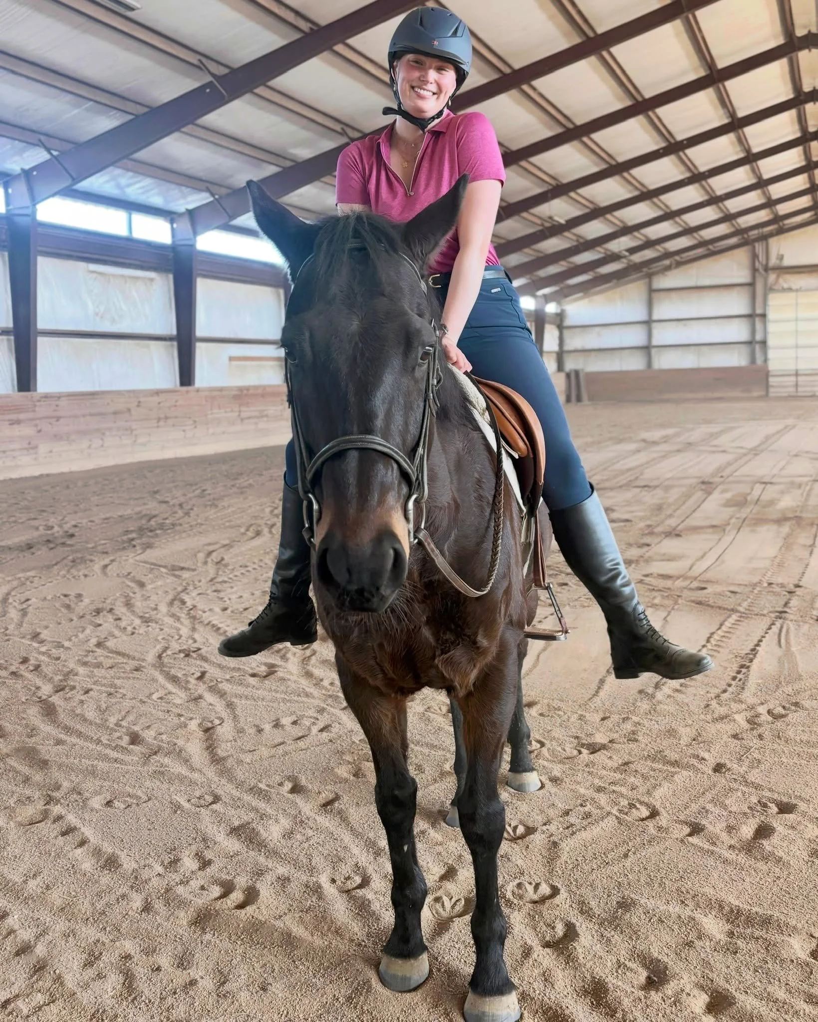 A woman wearing a helmet riding a dark brown horse inside a covered riding arena with sandy ground.