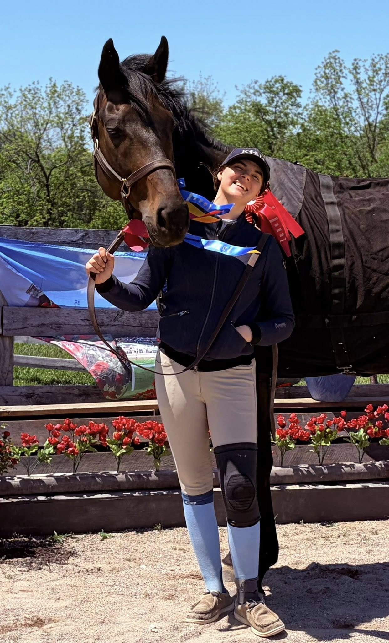 Young woman in equestrian riding outfit standing with a horse, holding the horse's bridle, outdoors on a sunny day with trees in the background and red flowers in the foreground.