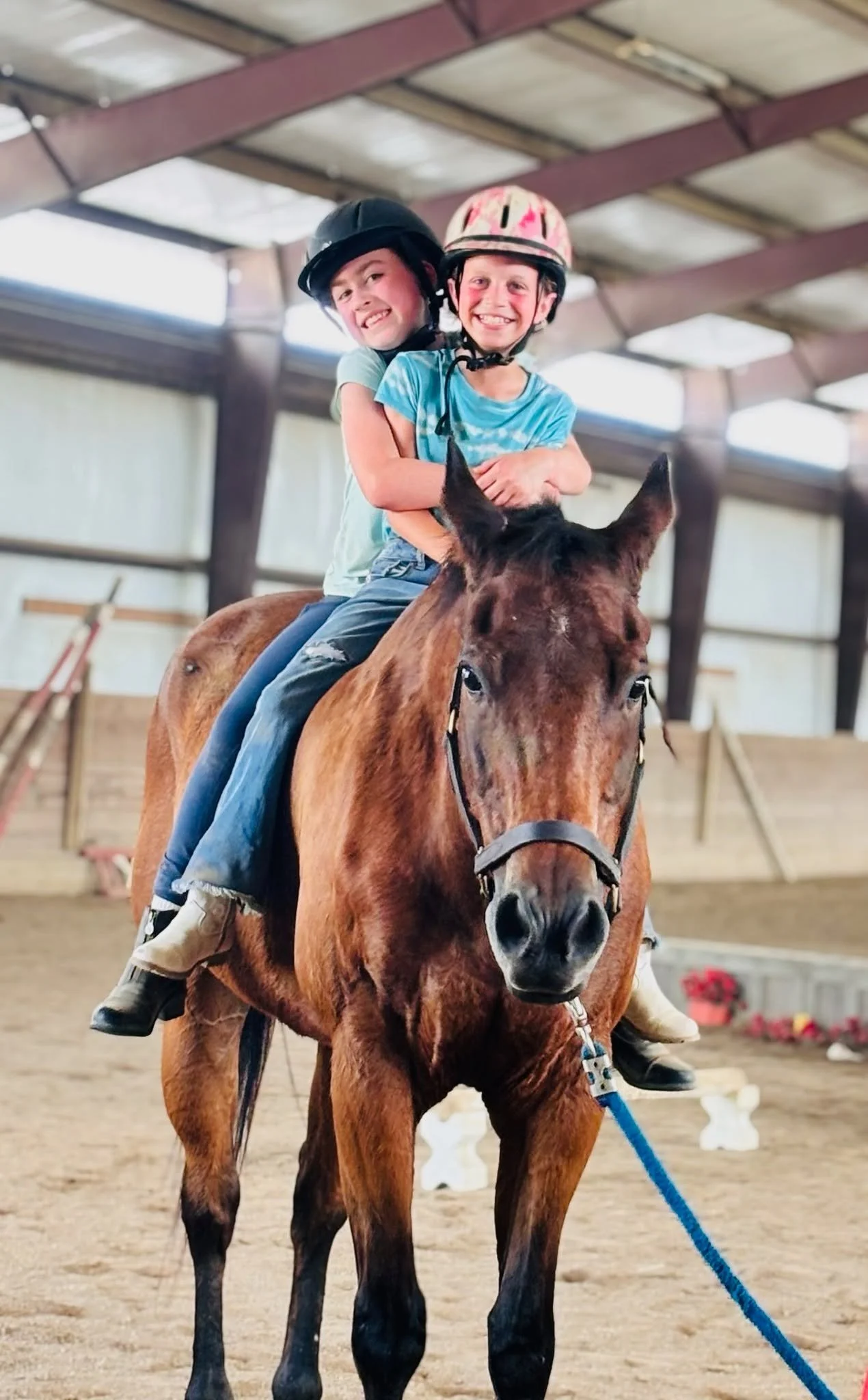 Two children riding a brown horse inside an indoor riding arena, wearing protective helmets and smiling.