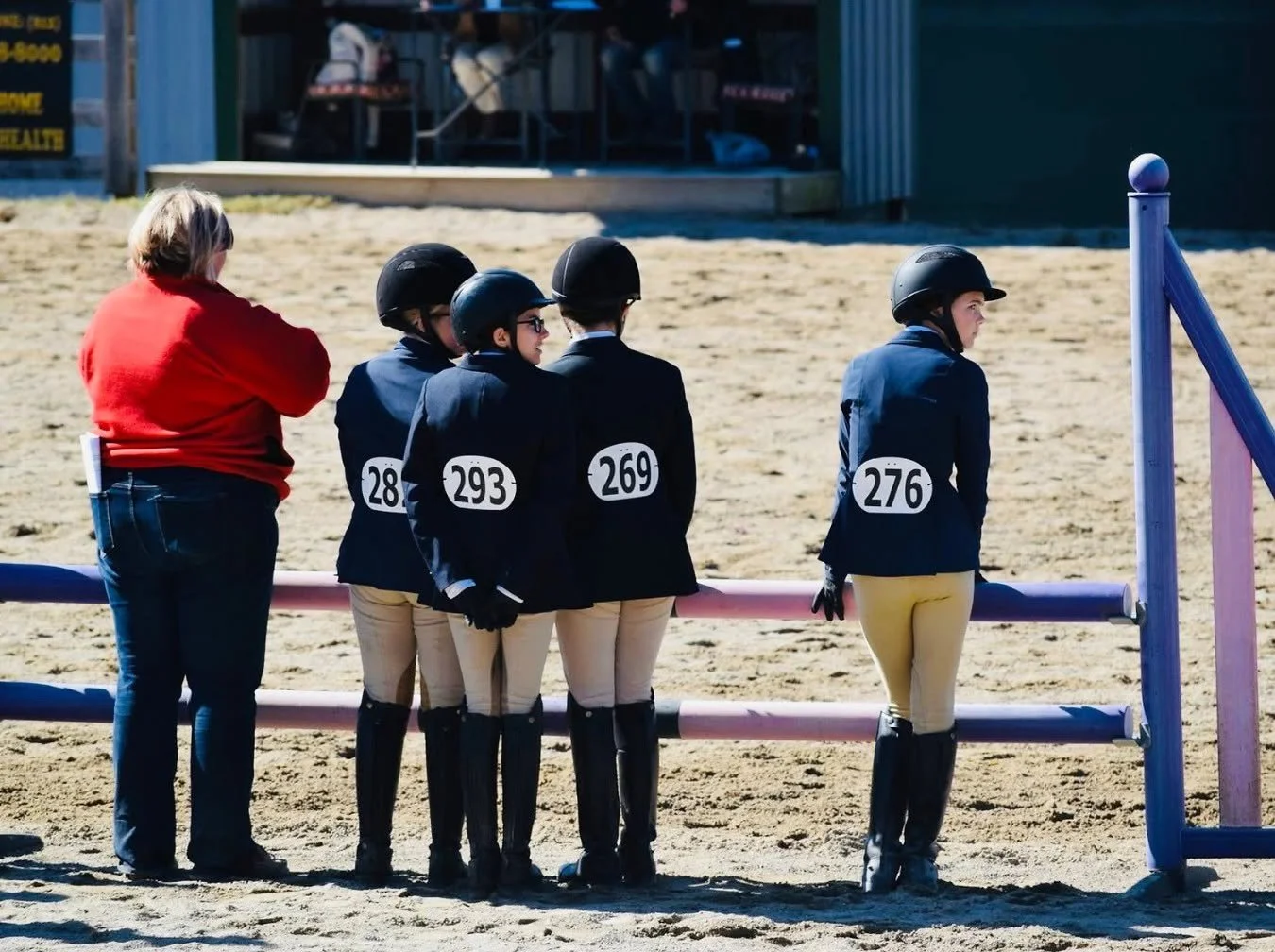 Four young equestrians in riding gear and helmets, standing in line at an outdoor horse-riding arena, with a woman in a red jacket standing nearby.