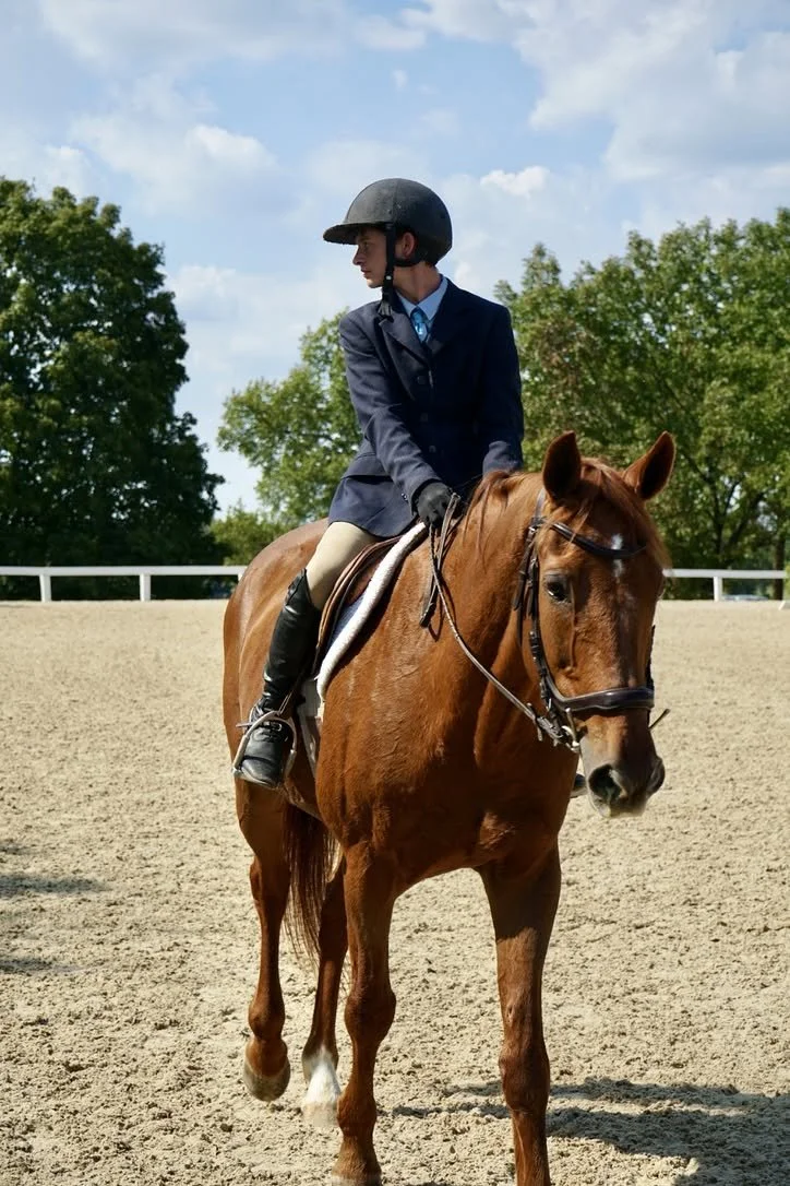 A person dressed in formal riding attire, including a helmet, blazer, and riding boots, is sitting on a chestnut horse in an outdoor riding arena surrounded by trees, with a partly cloudy sky overhead.