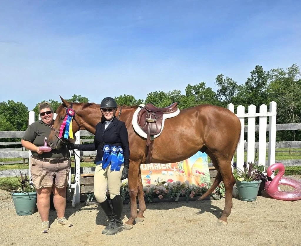Two smiling women and a horse with a pink and blue rosette ribbon, standing in a riding arena with a fenced background, potted plants, and pink flamingo pool floaties.