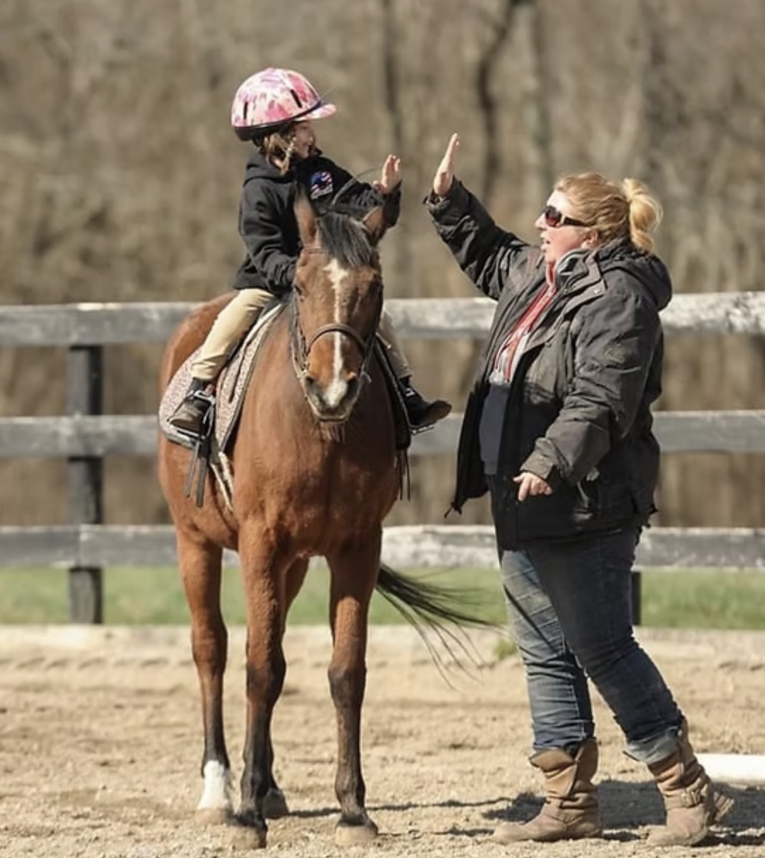 A young girl on horseback giving a high five to a woman standing outside in an outdoor riding arena during daytime.