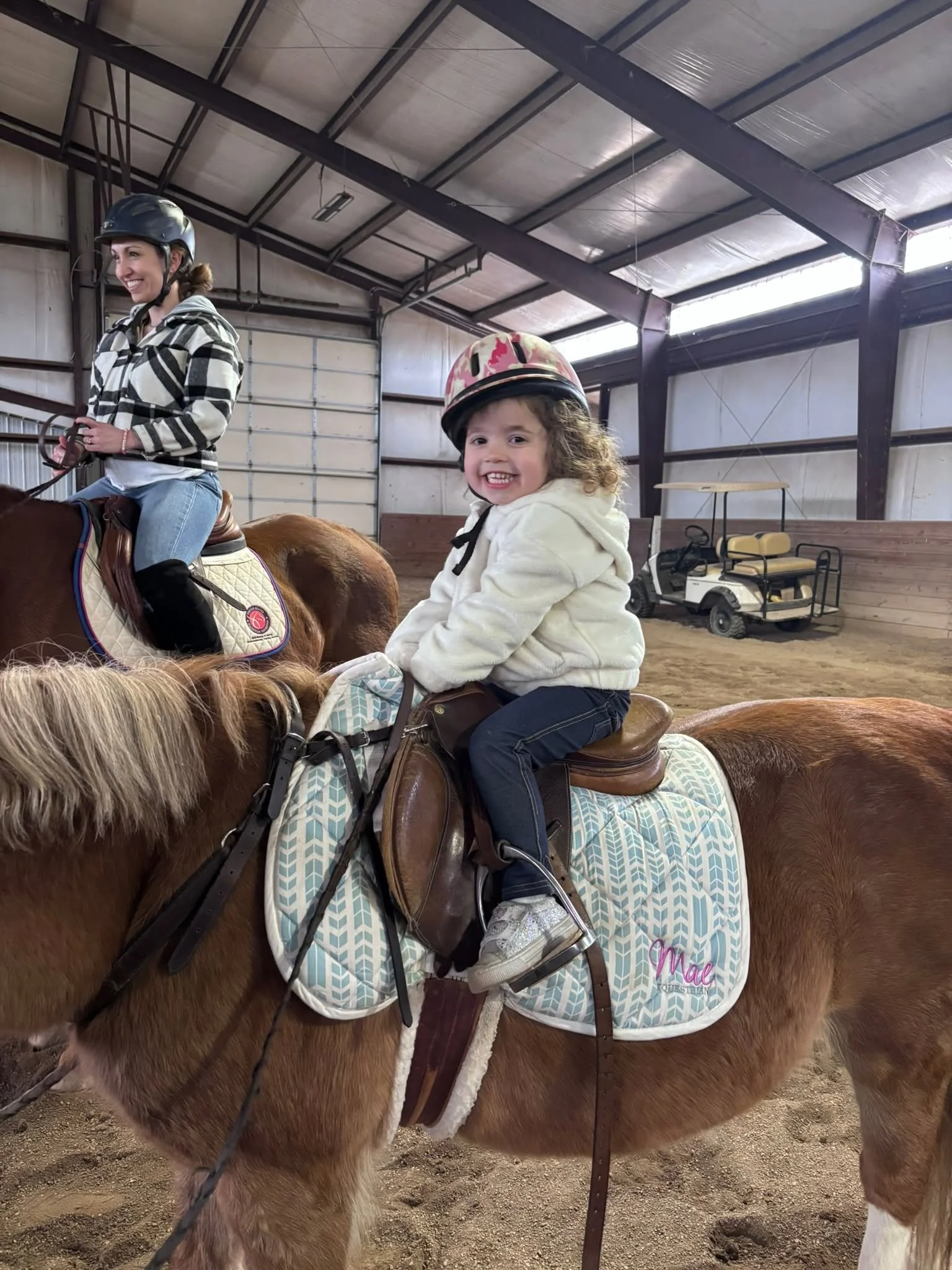 A young girl with curly hair, wearing a helmet and a white hoodie, riding a horse inside an indoor riding arena with a smiling woman on horseback nearby.