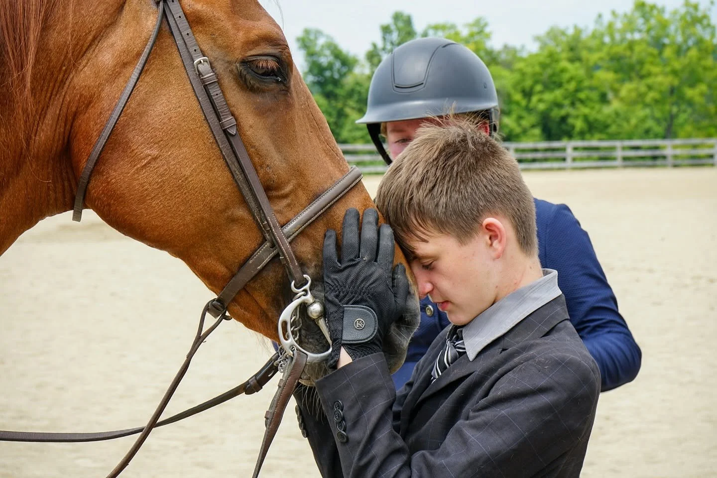 A young boy in formal attire with his eyes closed, gently pressing his forehead against a brown horse's nose. An adult wearing a helmet and a suit stands behind them, holding the horse's bridle in an outdoor riding area with green trees in the background.