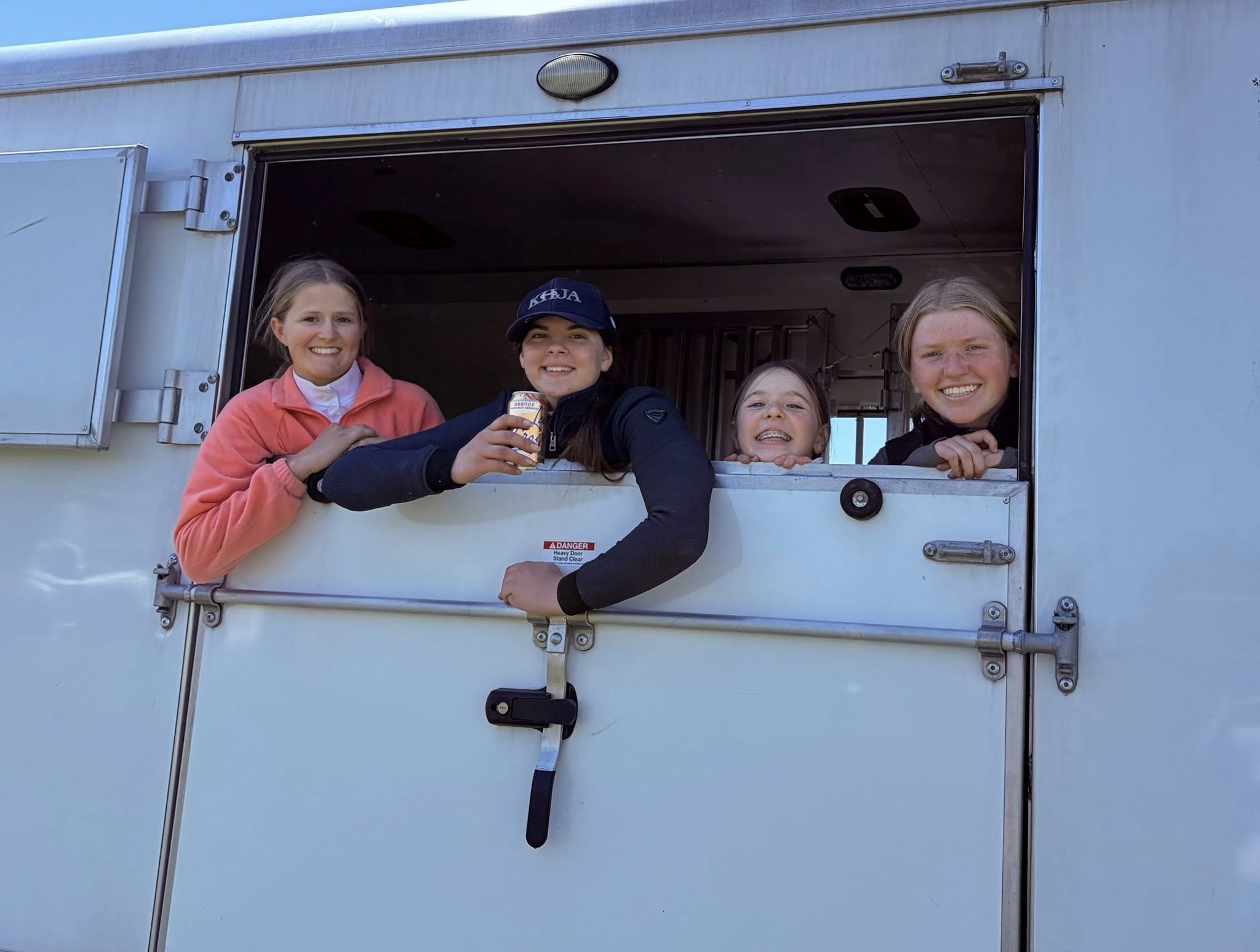 Four girls smiling and looking out of a service window, one girl holds a canned beverage, inside a vehicle or trailer.