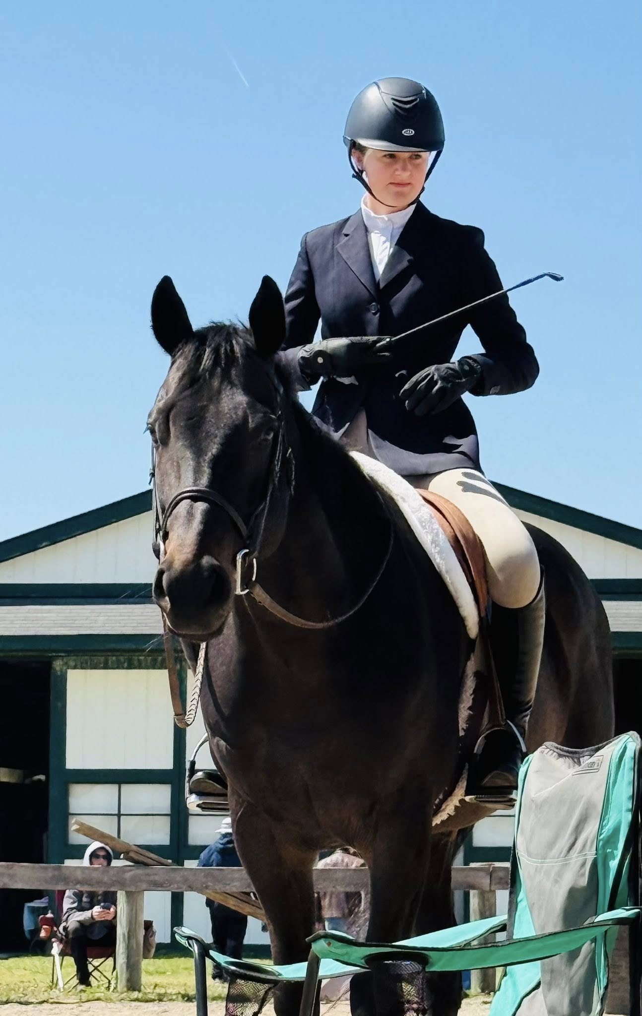 A young woman wearing a black helmet and riding a dark brown horse with a white stripe on its nose during an outdoor event. Several people are sitting or standing in the background, with blue sky above.