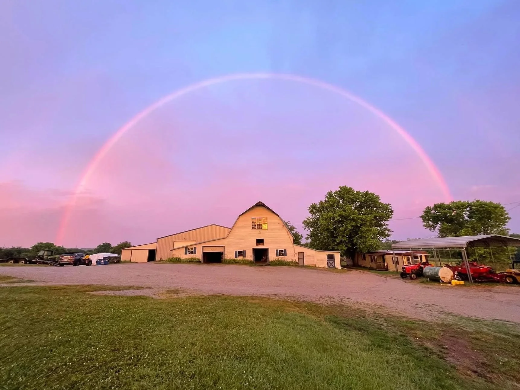 A farm scene at sunset with a large barn, trees, parked vehicles, and a rainbow arching across a pastel sky.