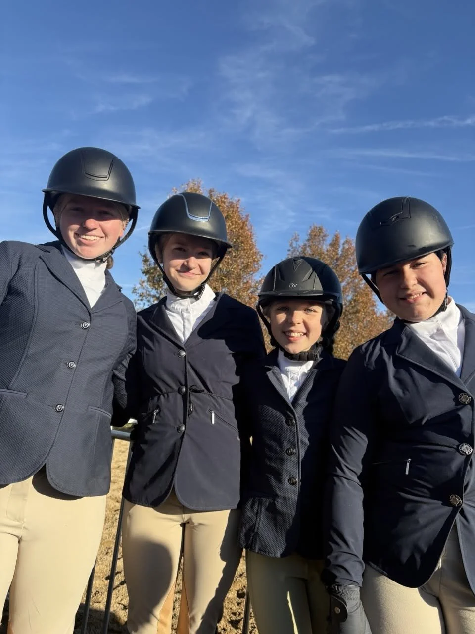 Four young women wearing equestrian riding helmets and navy blazers standing outdoors on a sunny day, with trees and a blue sky in the background.