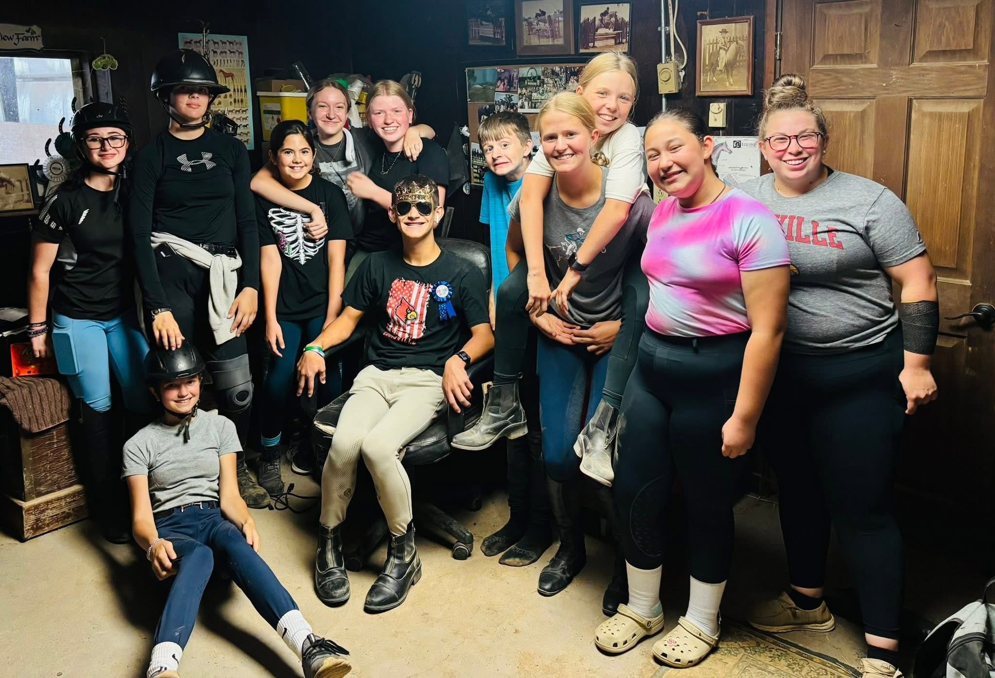 A group of children and teenagers gathered in a room, some wearing equestrian helmets and boots, smiling and posing for a photo.