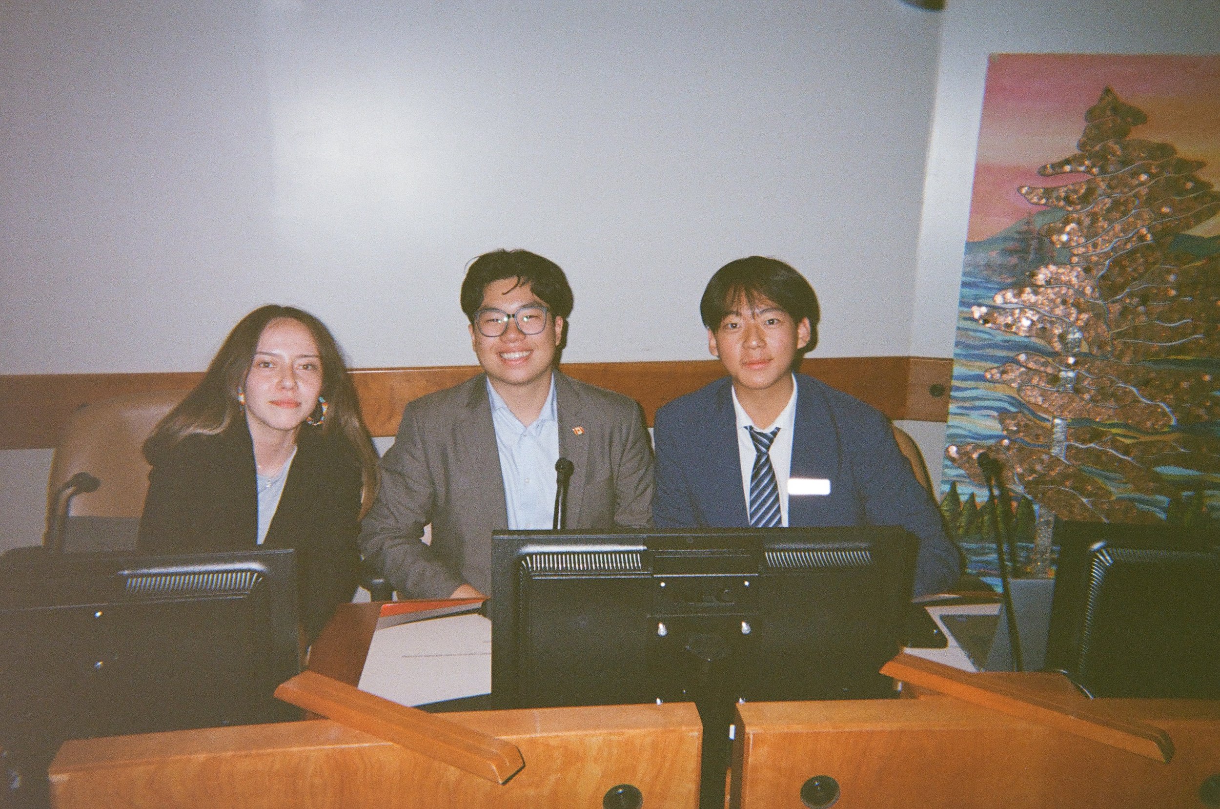 Three young adults sitting at a conference table with computers, in a room with a decorative painting of trees on the wall behind them.