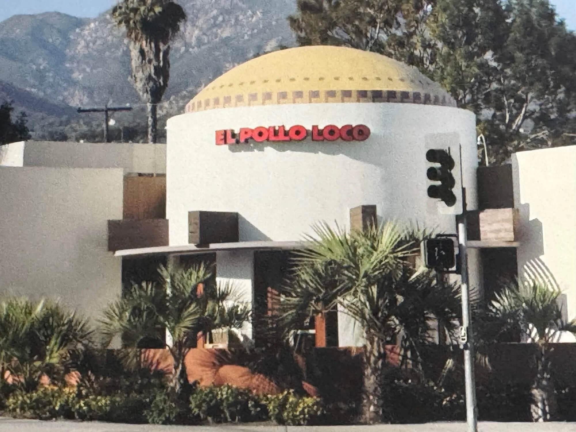 A white building with a rounded roof and red sign that reads 'El Pollo Loco' is surrounded by trees and mountains in the background.