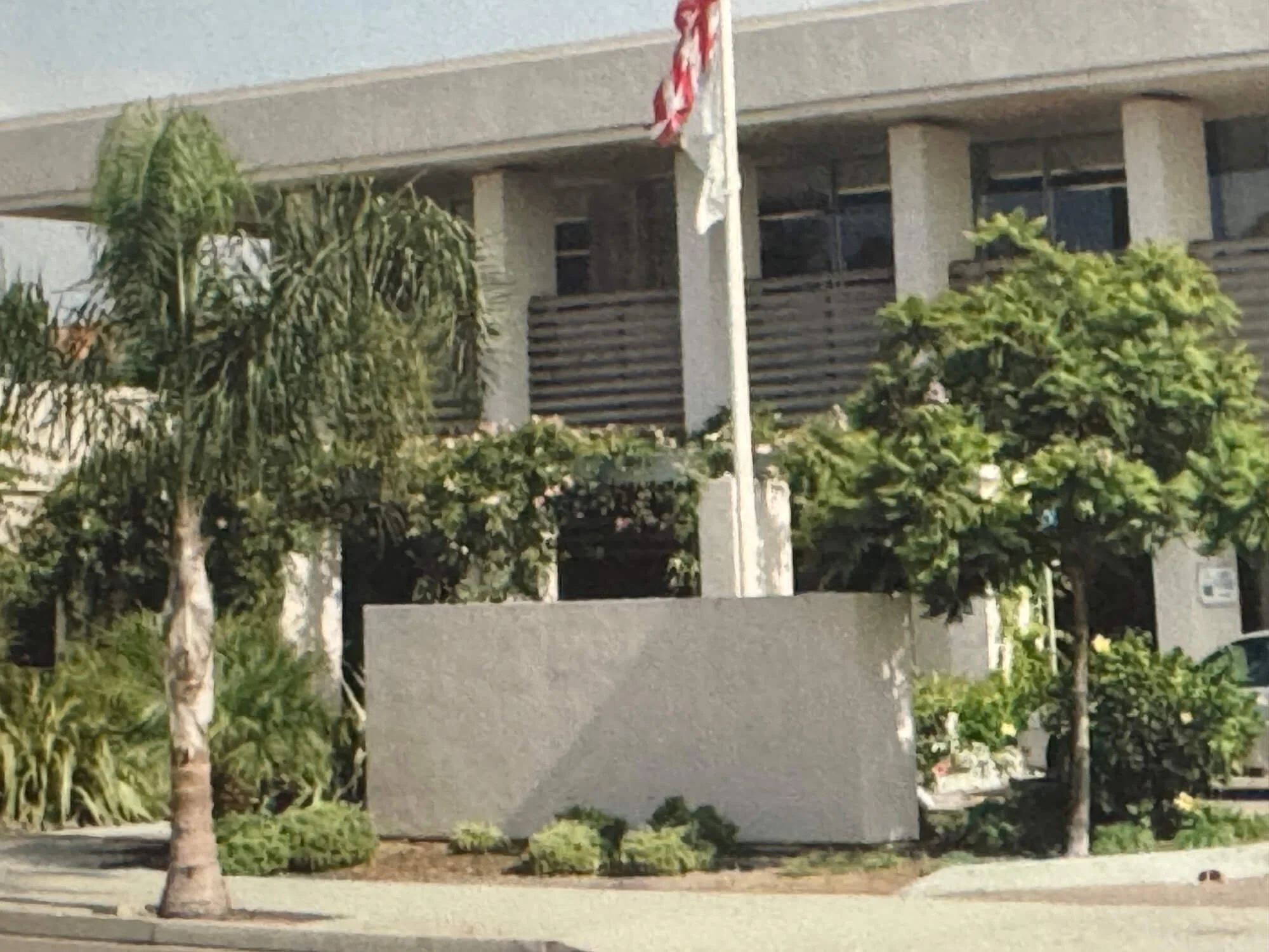 A modern residential building with a concrete facade and horizontal wooden slats covering the windows. There are green trees and shrubs in front of the building and a sidewalk along the street.