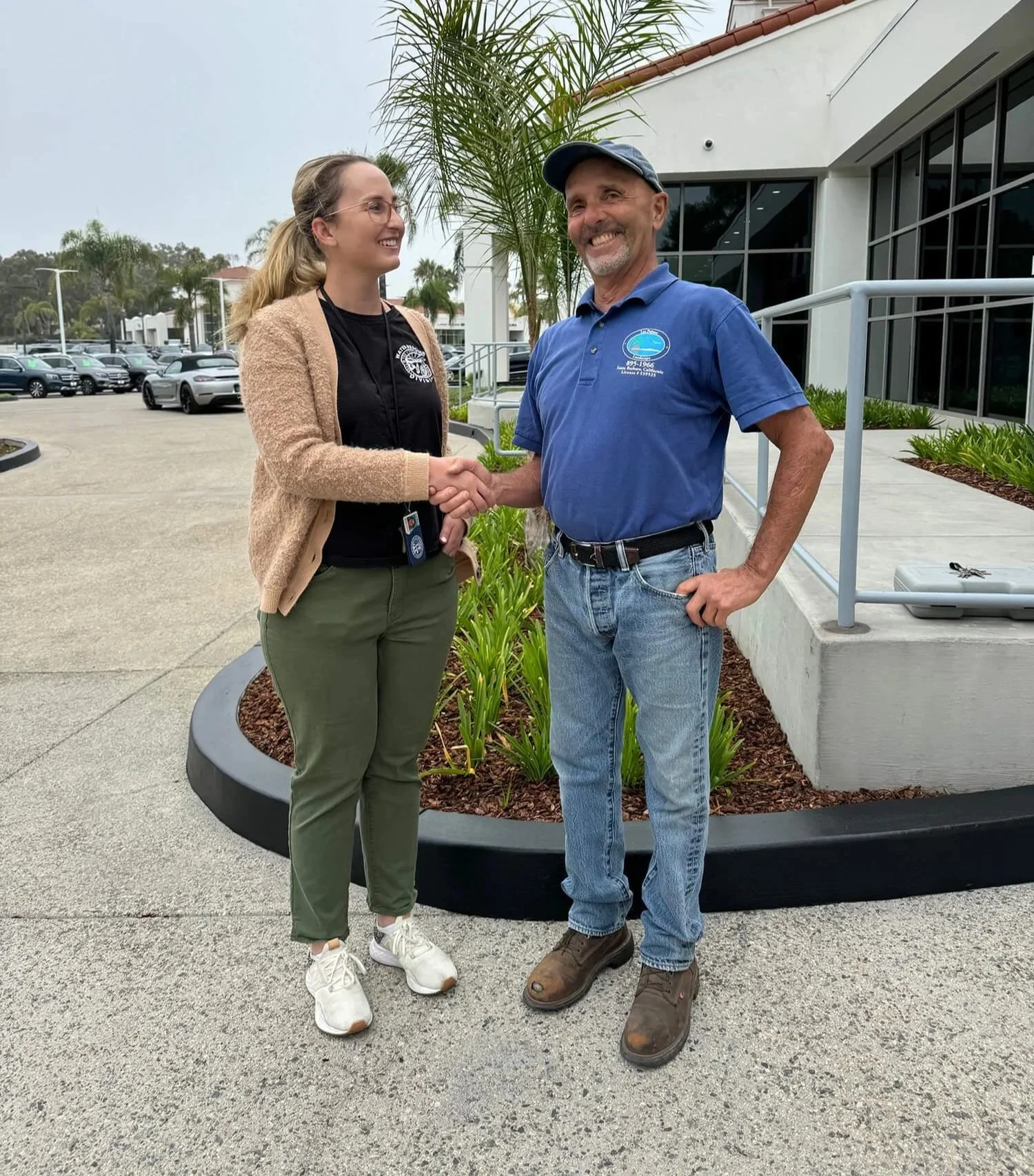 A woman and a man are shaking hands outside a building. The woman has glasses, long blonde hair, a black shirt, beige cardigan, and green pants. The man is wearing a blue polo shirt, jeans, and brown work boots, and is smiling. There are palm trees, parked cars, and a white building with large windows in the background.