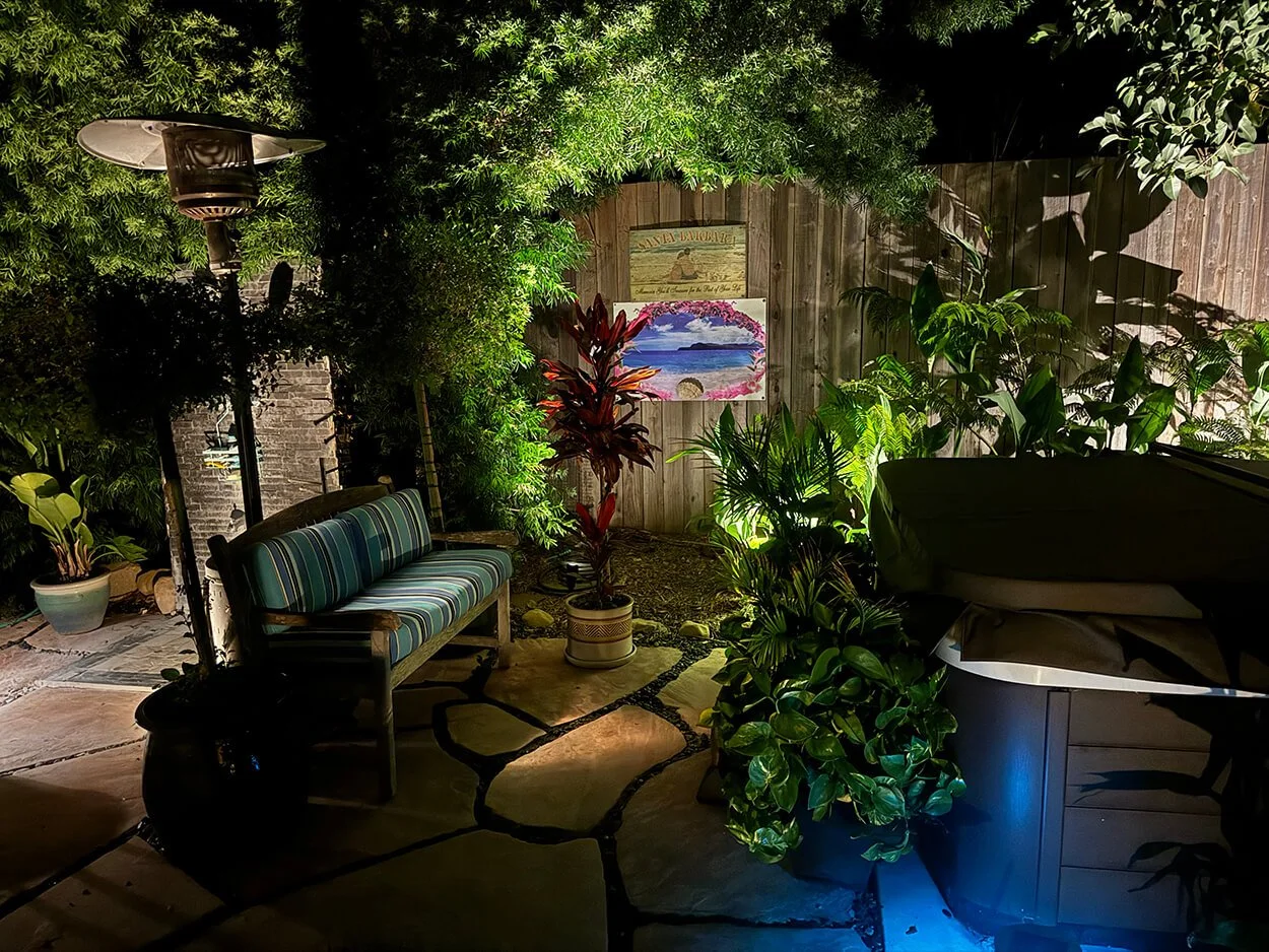 Nighttime backyard patio with outdoor heater, striped bench, lush green plants, wooden fence, and colorful paintings on the wall.