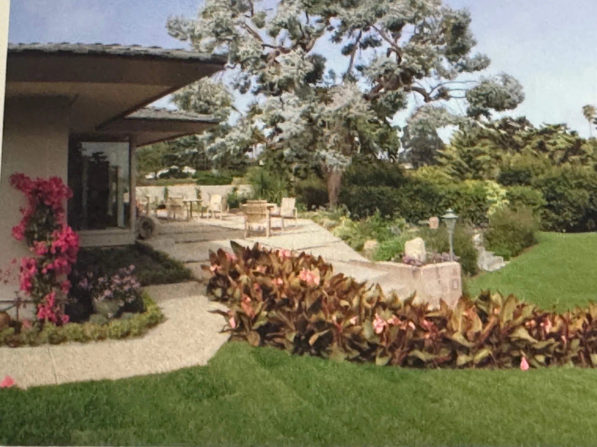A house with a porch decorated with pink flowers, and a garden with green grass, leafy shrubs, and trees, including a large pine tree in the background, under a clear sky.