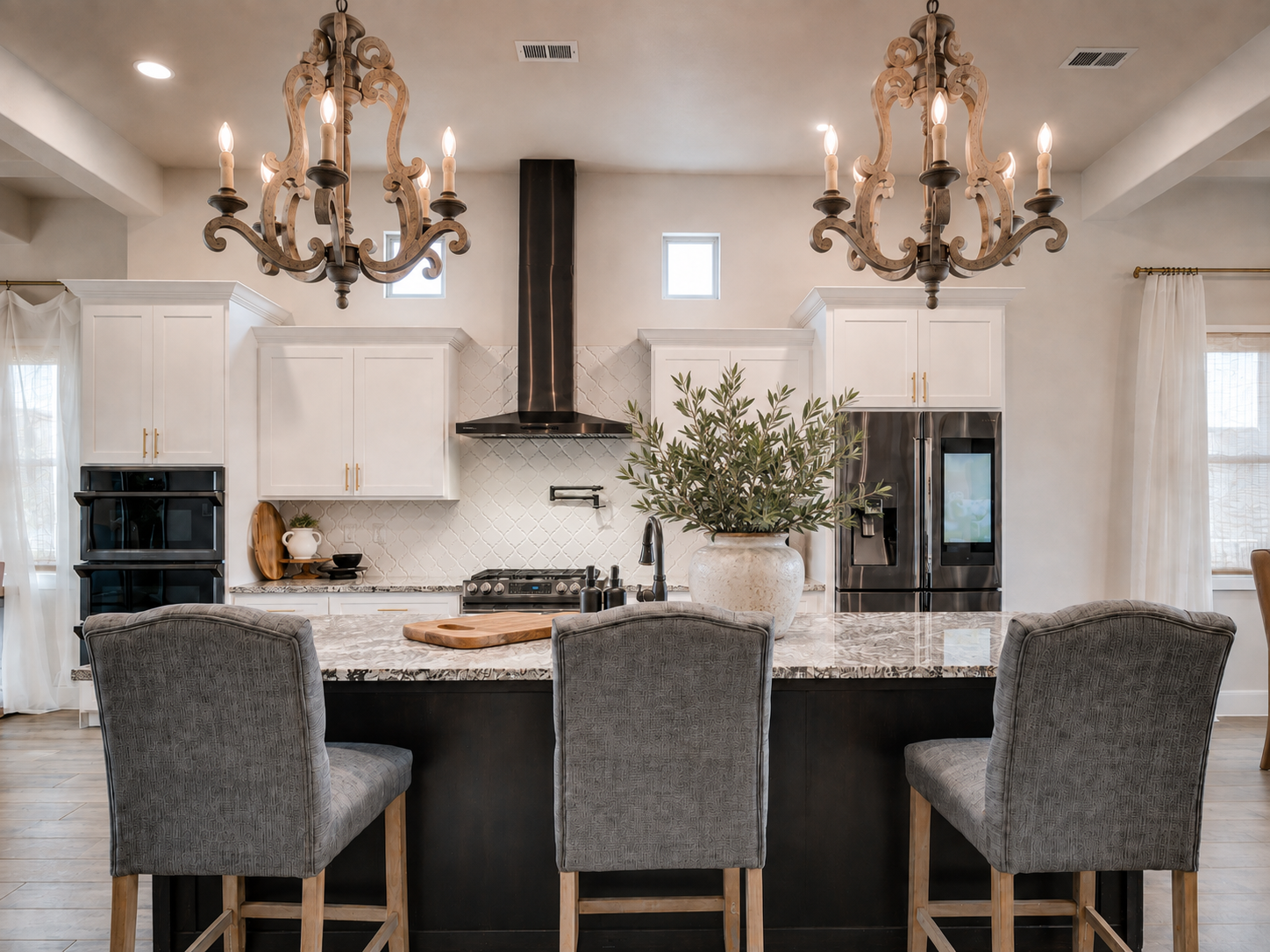 Crisp white cabinetry and a rich espresso island create a grounded contrast, softened by warm wood chandeliers, brass details, sculptural greenery, and textured white tile.