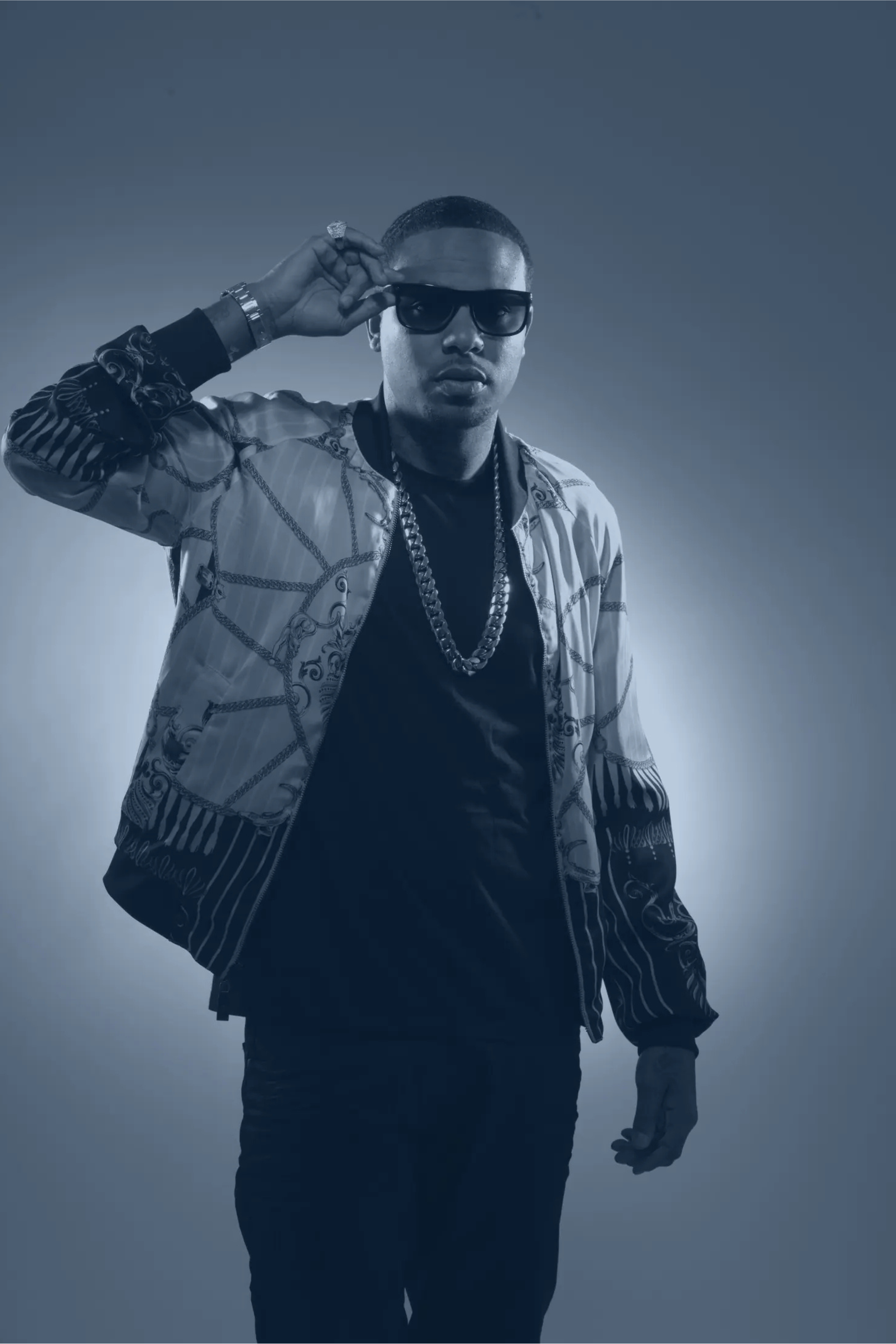 A man wearing dark sunglasses, a patterned jacket, a black shirt, and multiple gold chains, adjusting his sunglasses with his right hand, against a plain background.