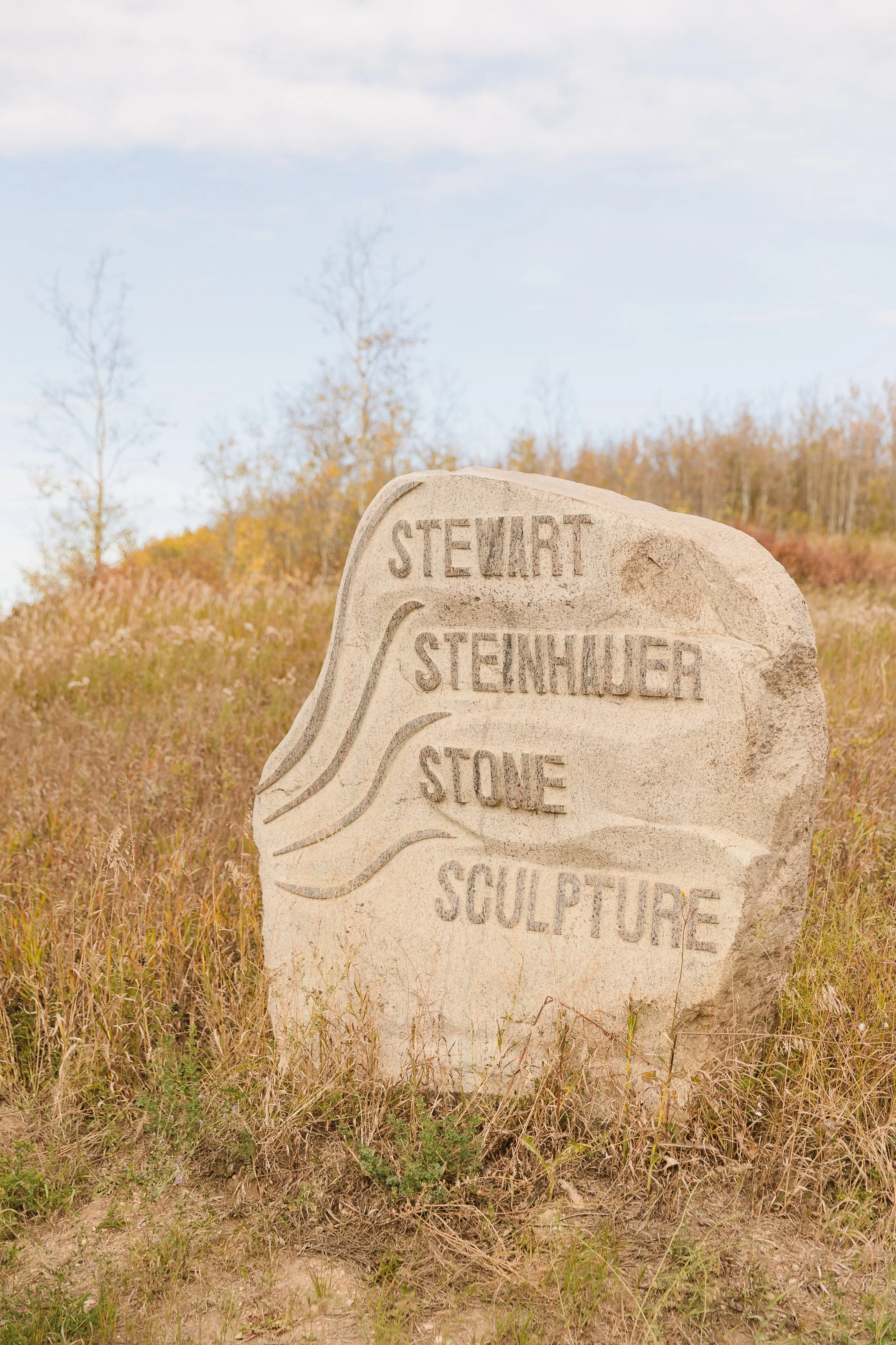 A large stone sculpture with the words 'Stewart', 'Steinhauer', 'Stone', and 'Sculpture' carved into it, situated outdoors in a grassy field with trees and a blue sky in the background.