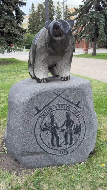 A polished granite bear statue on a large stone base with an engraving of two Native Americans shaking hands, the text 'Indian Treaty No. 6 1875', and crossed rifles, situated outdoors among trees and grass.