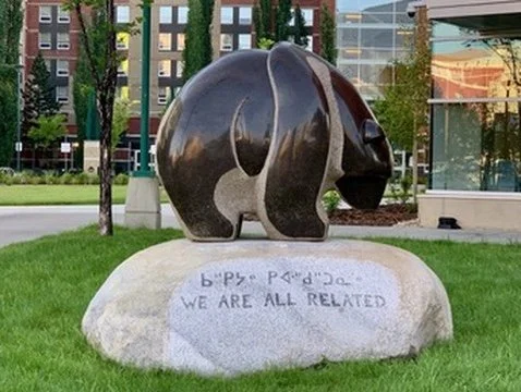 Bronze bear sculpture on a large gray rock with engraved inscription, set on grass in an urban park.
