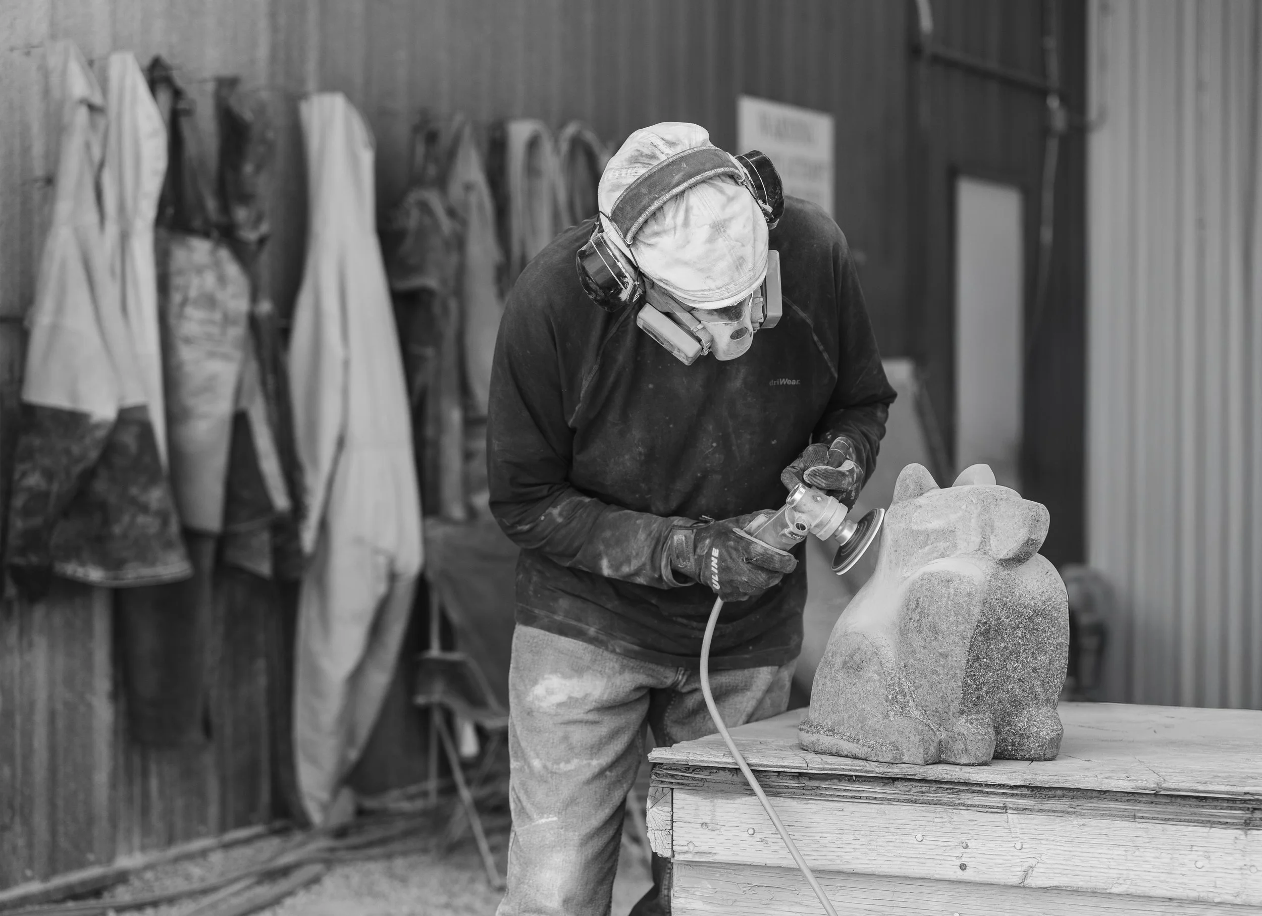 A person wearing a protective mask and goggles working on a stone sculpture in a workshop, with aprons hanging on the wall in the background.