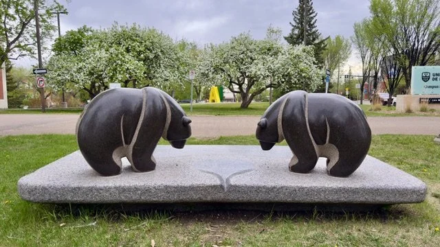 Sculpture of two bears facing each other, carved from dark stone, mounted on a granite base, in a park with blossoming trees and a corporate sign in the background.