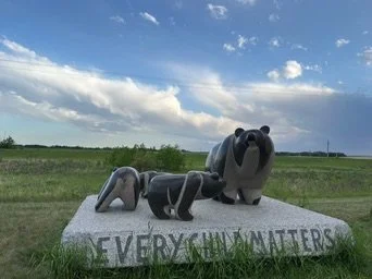 Outdoor sculpture of three bears on a concrete base with 'EVERYONE MATTERS' written on it, set in a grassy field under a partly cloudy sky.