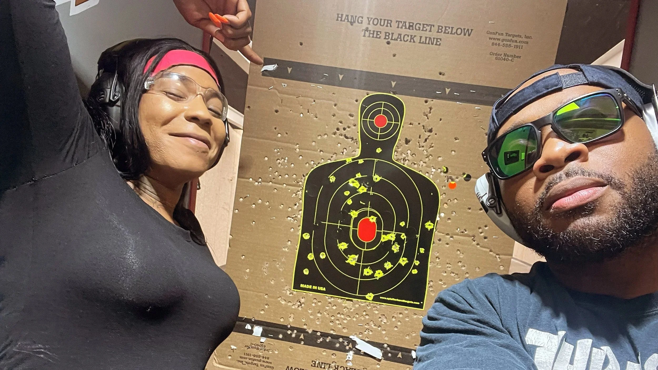 A woman and man at a shooting range taking a selfie, standing next to a cardboard target with bullet holes. The woman wears glasses, a pink headband, and safety headphones, smiling with her eyes closed. The man wears sunglasses, a cap, and safety glasses, looking at the camera. The target has a black silhouette with red and yellow markings, and a printed message that reads, "HANG YOUR TARGET BELOW THE BLACK LINE."