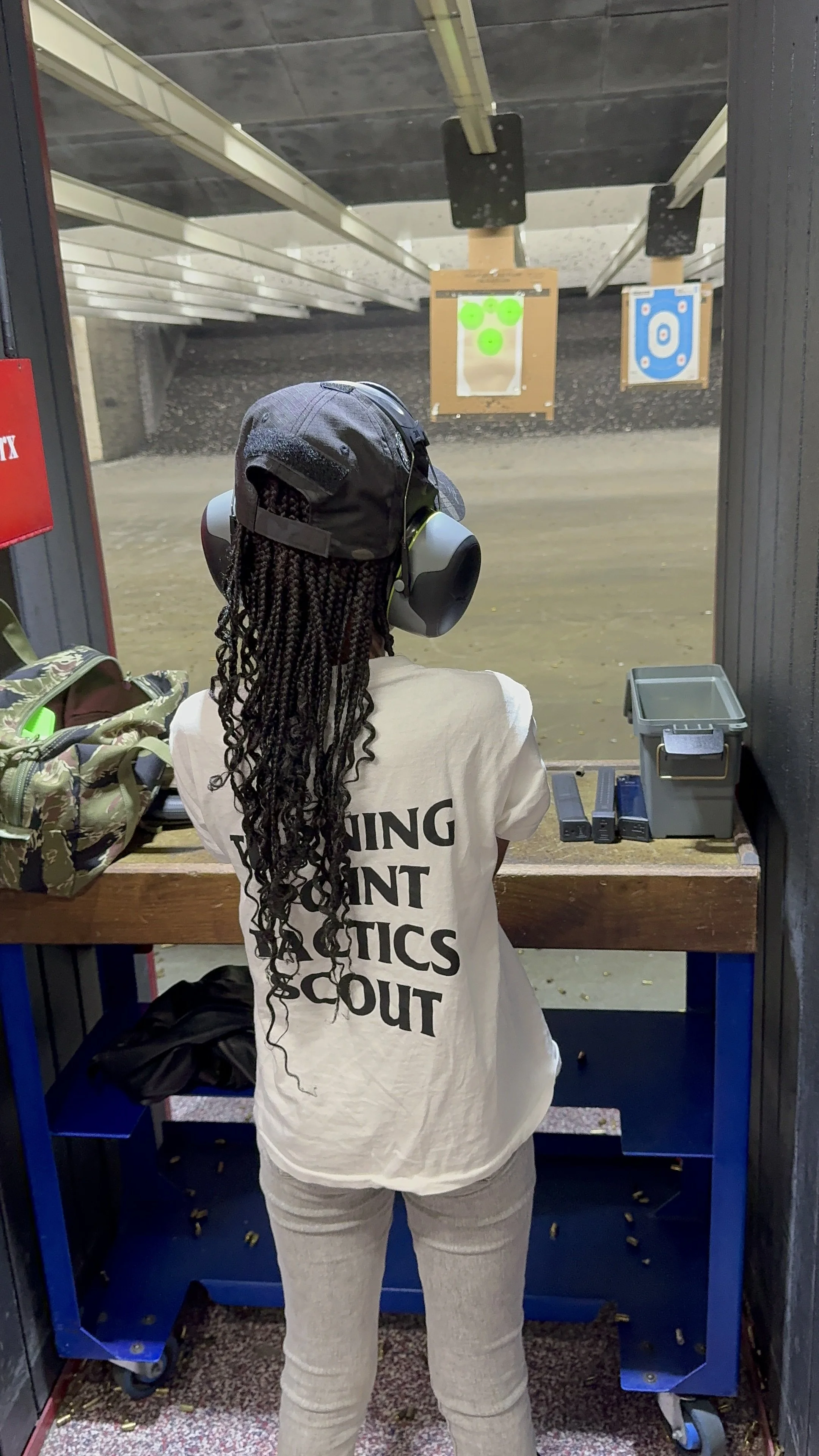 A female child at a shooting range wearing hearing protection and a cap, aiming at targets in the distance. She has long braids and is wearing a white shirt with black text on the back. There are firearms placed on the table in front of her.