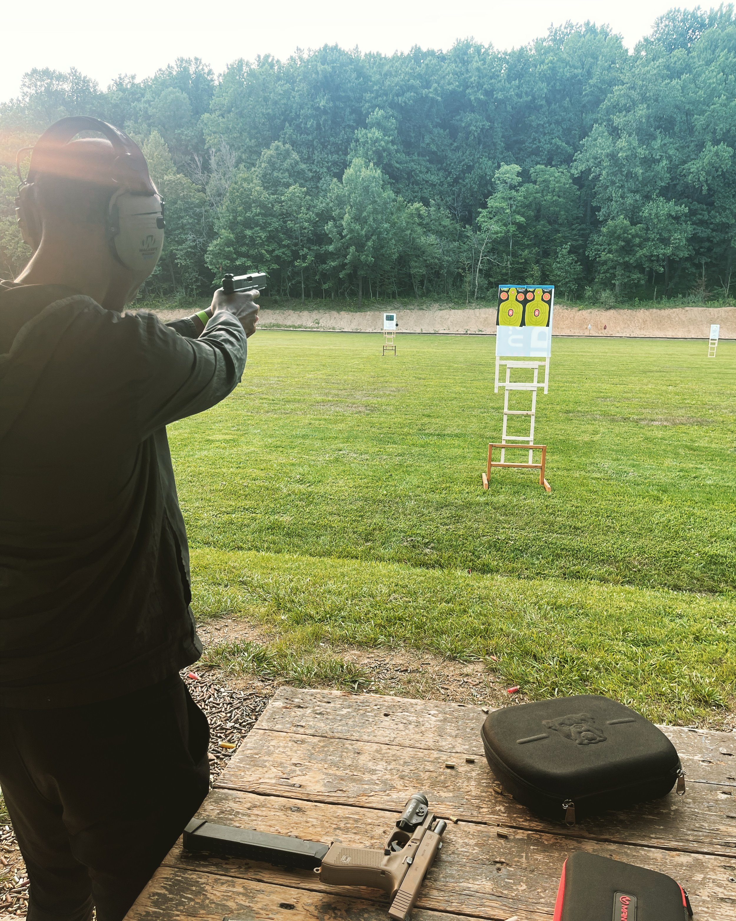 A person wearing hearing protection aims a handgun at outdoor shooting targets, with various firearm accessories and cases on a wooden table.