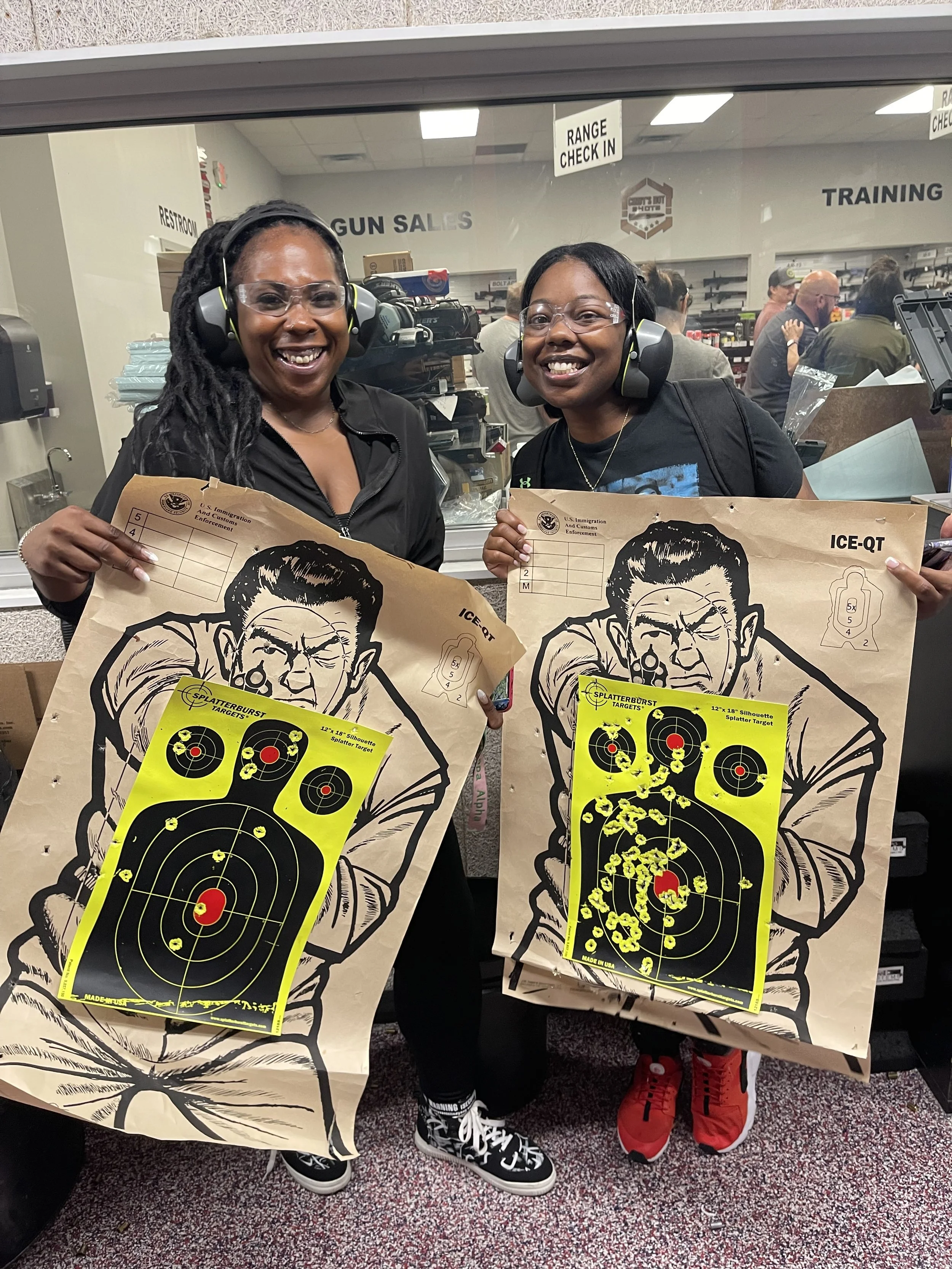 Two smiling women wearing safety glasses and ear protection hold shooting target sheets with bullet hole patterns, standing in front of a glass window at a gun range.