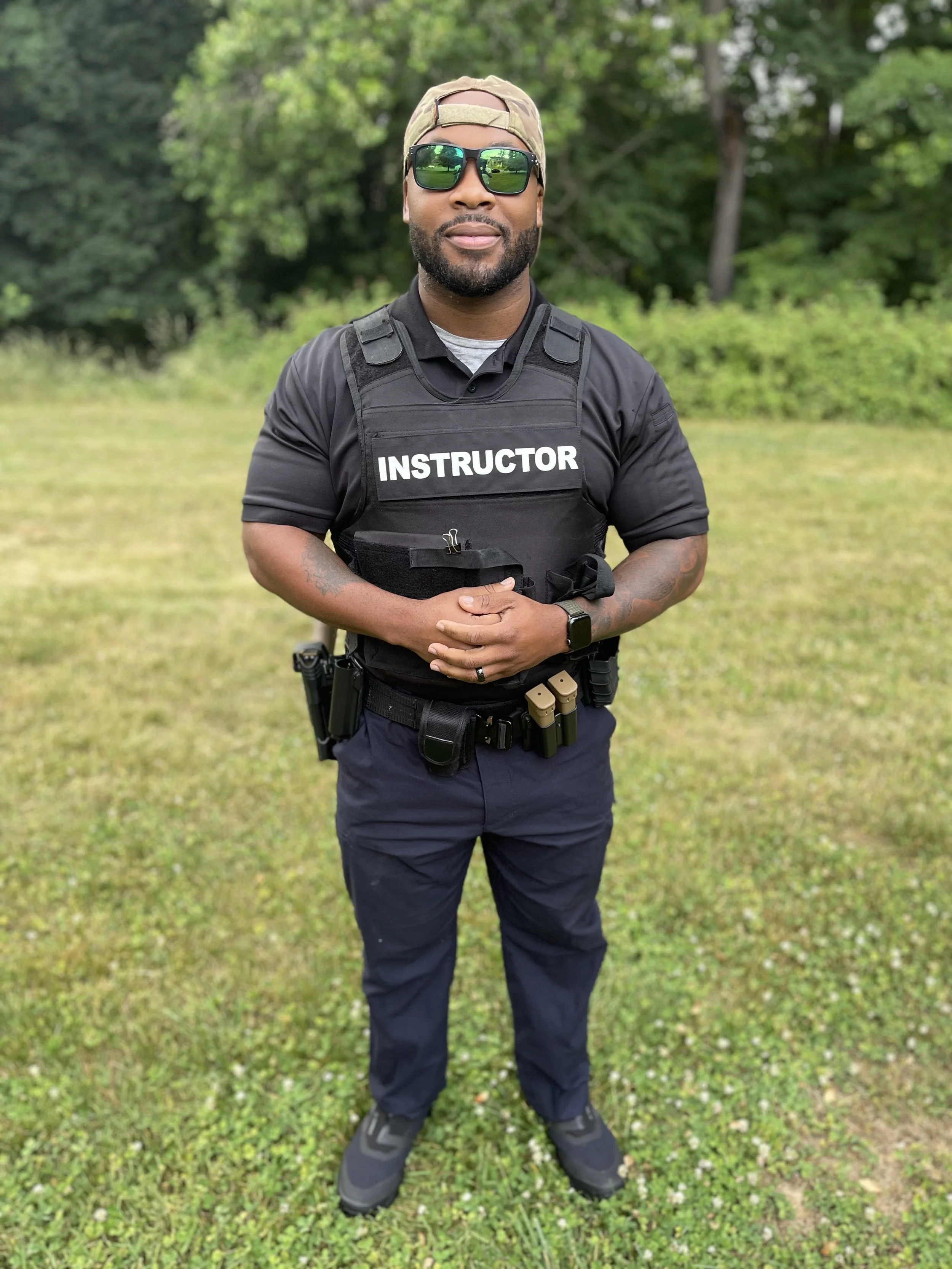 A man standing outdoors on grass, wearing sunglasses, a camo cap backwards, a black tactical vest labeled 'INSTRUCTOR', dark pants, and tactical gear, with a wooded background.