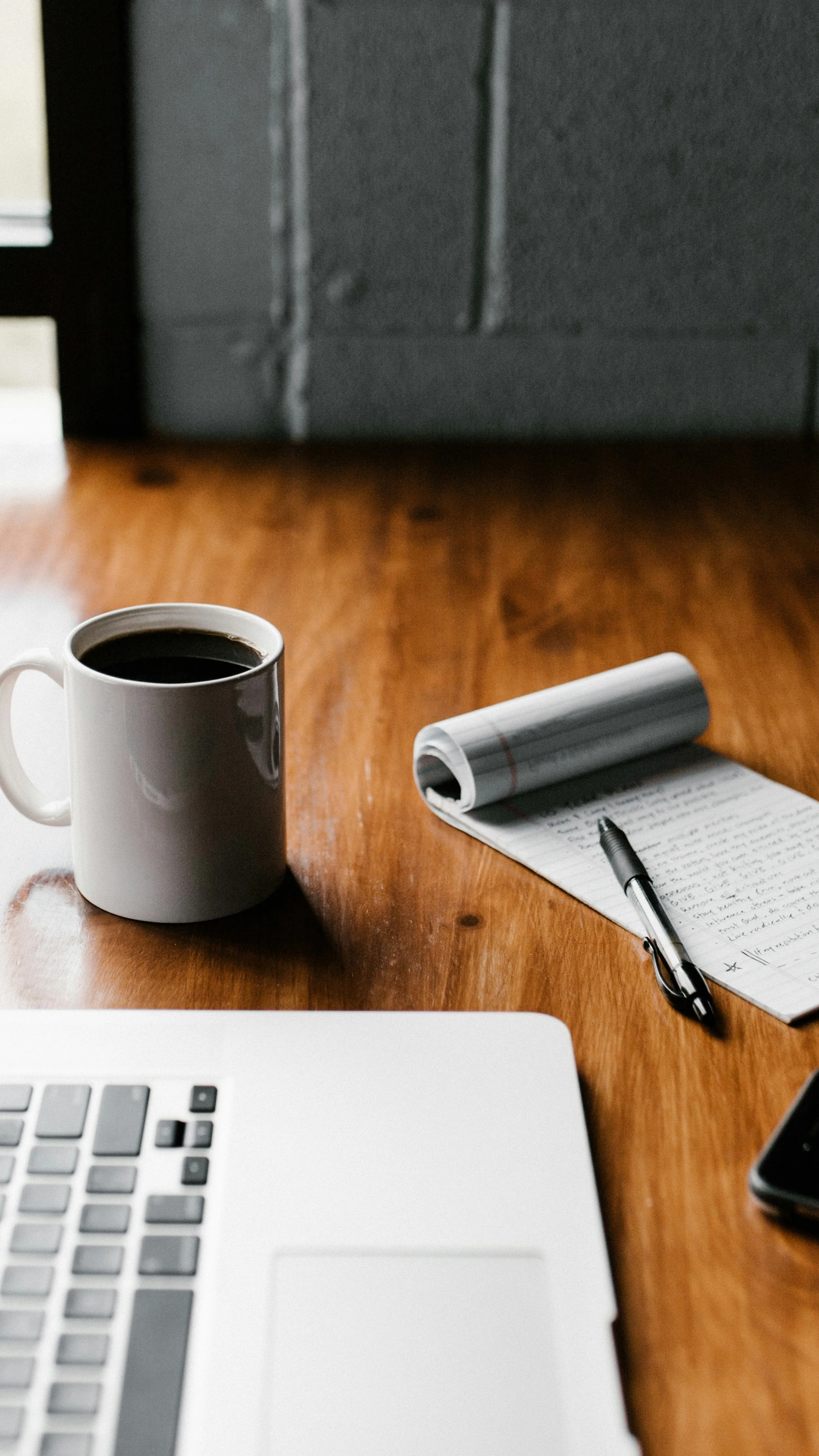 A wooden desk with a white mug of coffee, a notepad with handwritten notes, a black pen, and part of a silver laptop keyboard visible. Background shows a dark wall and hardwood floor.